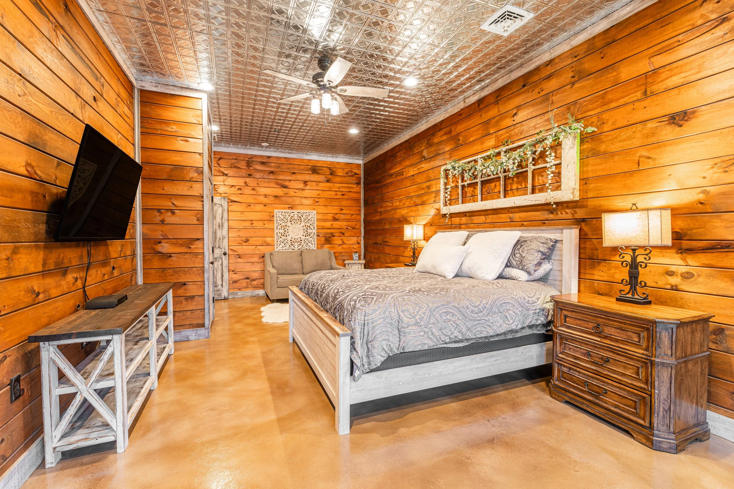 A bedroom with walls made of wooden planks, a bed with patterned bedding, a bedside table with a lamp, a beige sofa, a wall decor piece, and a window with a plant decoration, elevated ceiling with a ceiling fan, and a mounted flat-screen TV.