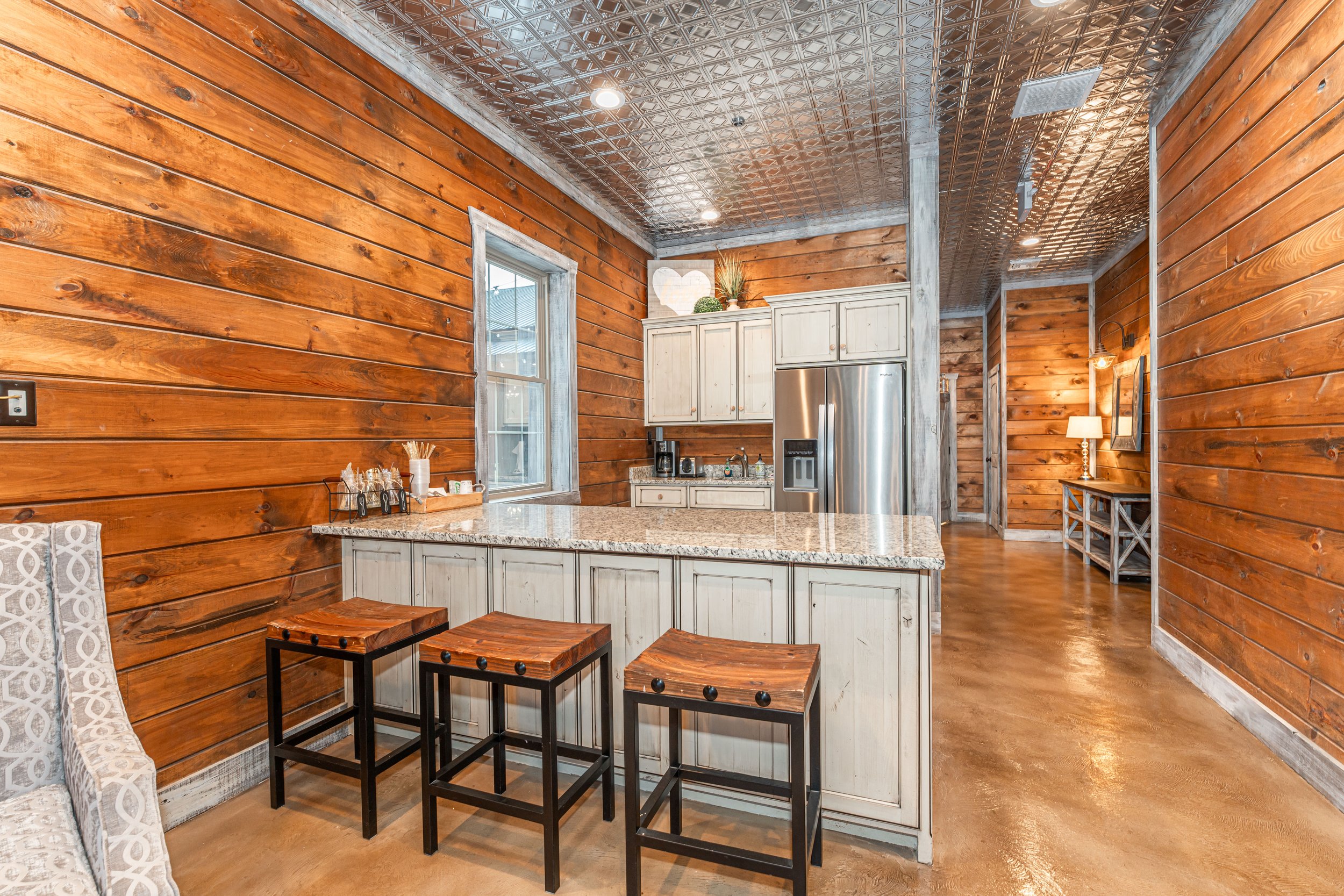 Interior view of a rustic kitchen with wood-paneled walls, granite countertop, stainless steel refrigerator, white cabinets, and decorative accents like a heart-shaped wall hanging and plants.