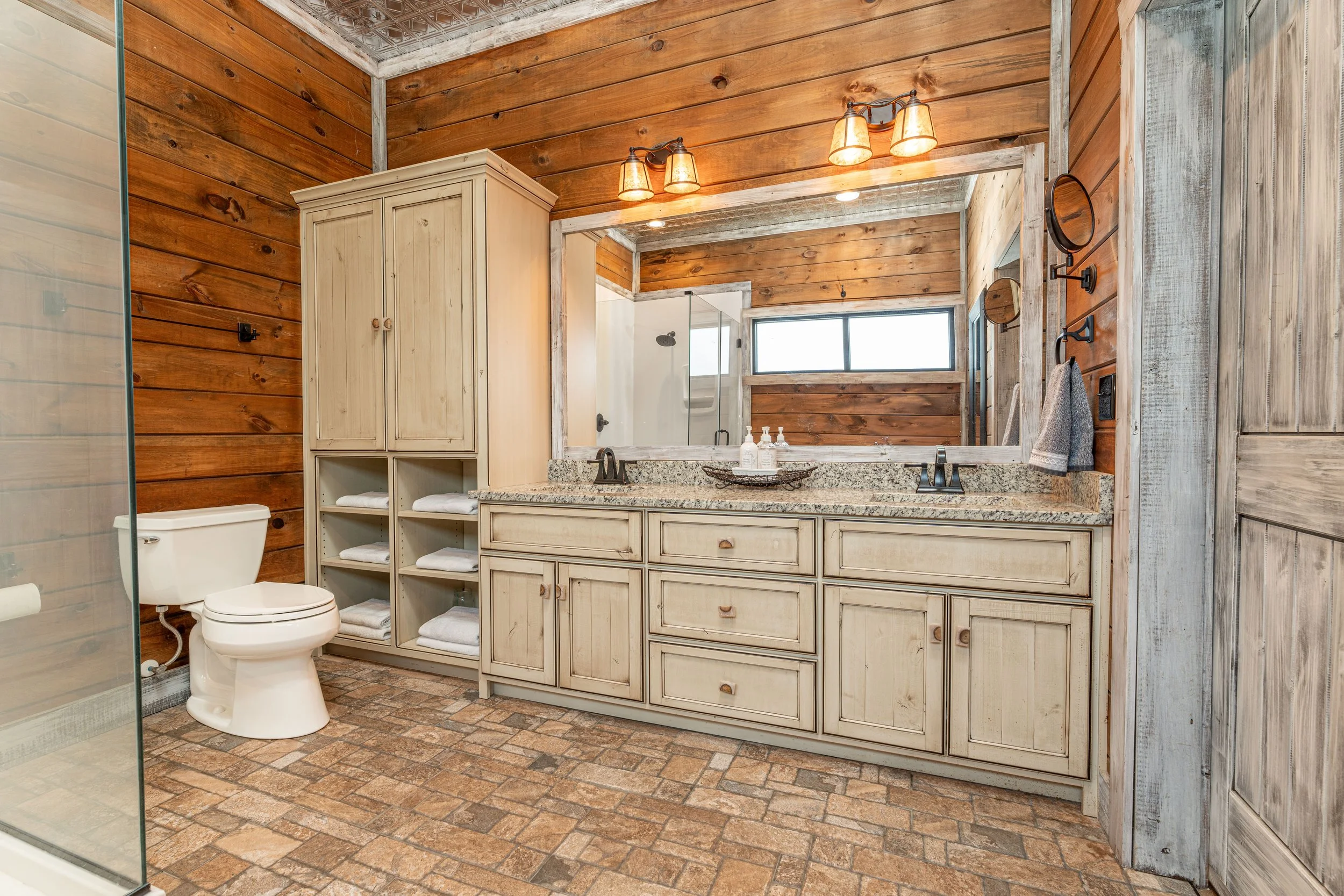 Bathroom with rustic design featuring wood-paneled walls, a large mirror above a granite countertop with double sinks, a wicker basket with soap, and a rustic wardrobe. There is a glass-enclosed shower area, exposed brick flooring, and a window provi