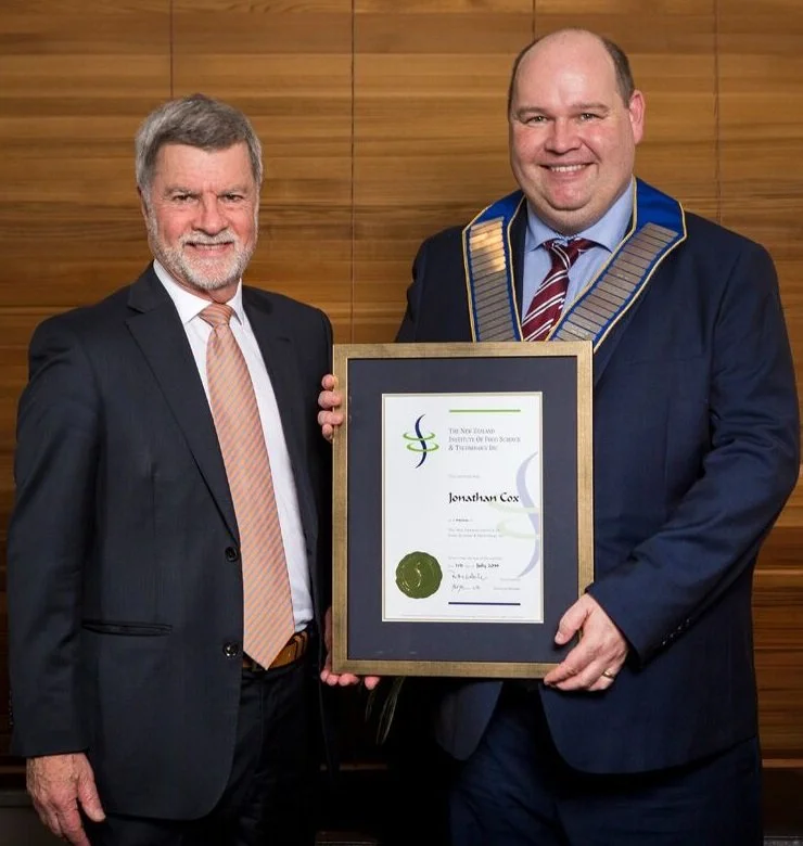 Two men in suits pose for a photo, one is holding a framed certificate. The man on the right is wearing a ceremonial chain of office, indicating a formal or official event. They are standing in front of a wooden wall.