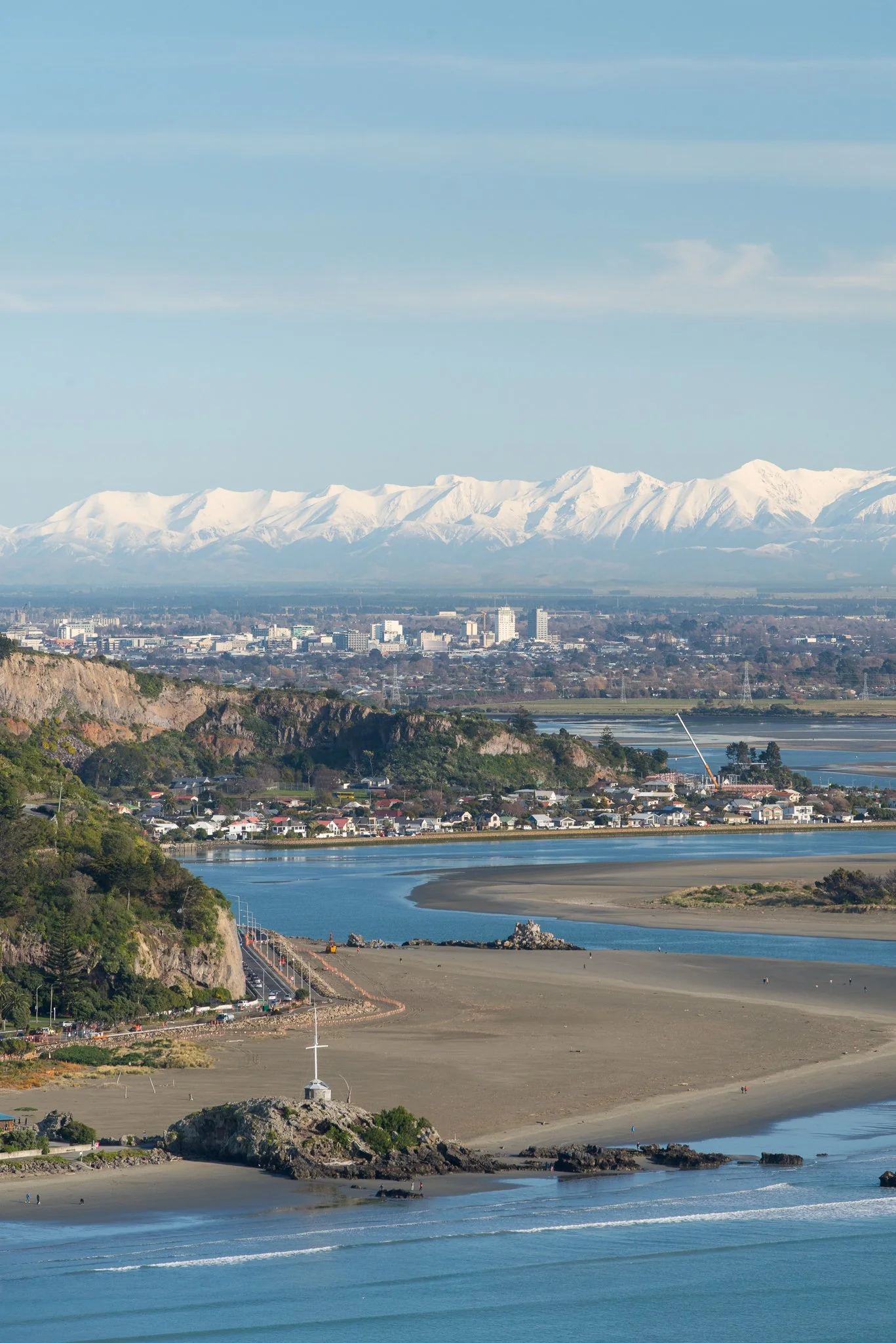 Scenic view of a coastal area with a beach, river, hillside, and distant snow-capped mountains under a partly cloudy sky.