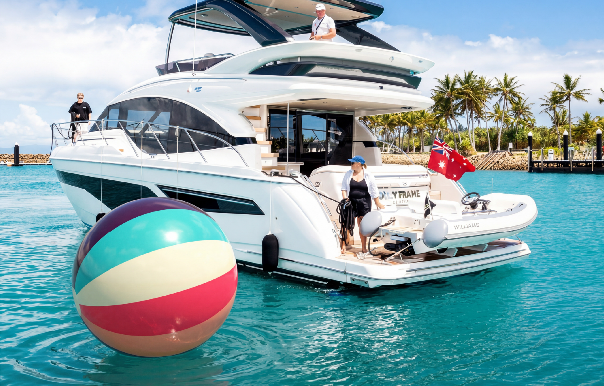 A large white yacht with two deck levels is floating in turquoise water near a tropical shoreline with palm trees. There is a person on the upper deck, and two women, one in a white hat and one in a blue cap, are on the lower deck. A colorful beach ball is in the foreground, and a boat with an Australian flag is docked at the stern.