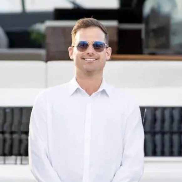 A smiling man wearing sunglasses and a white dress shirt, standing outdoors near a patio or pool area.