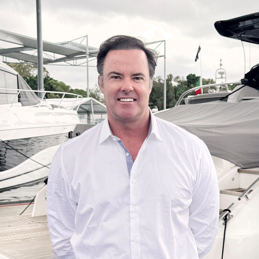 A smiling man in a white button-up shirt standing on a dock with yachts in the background.