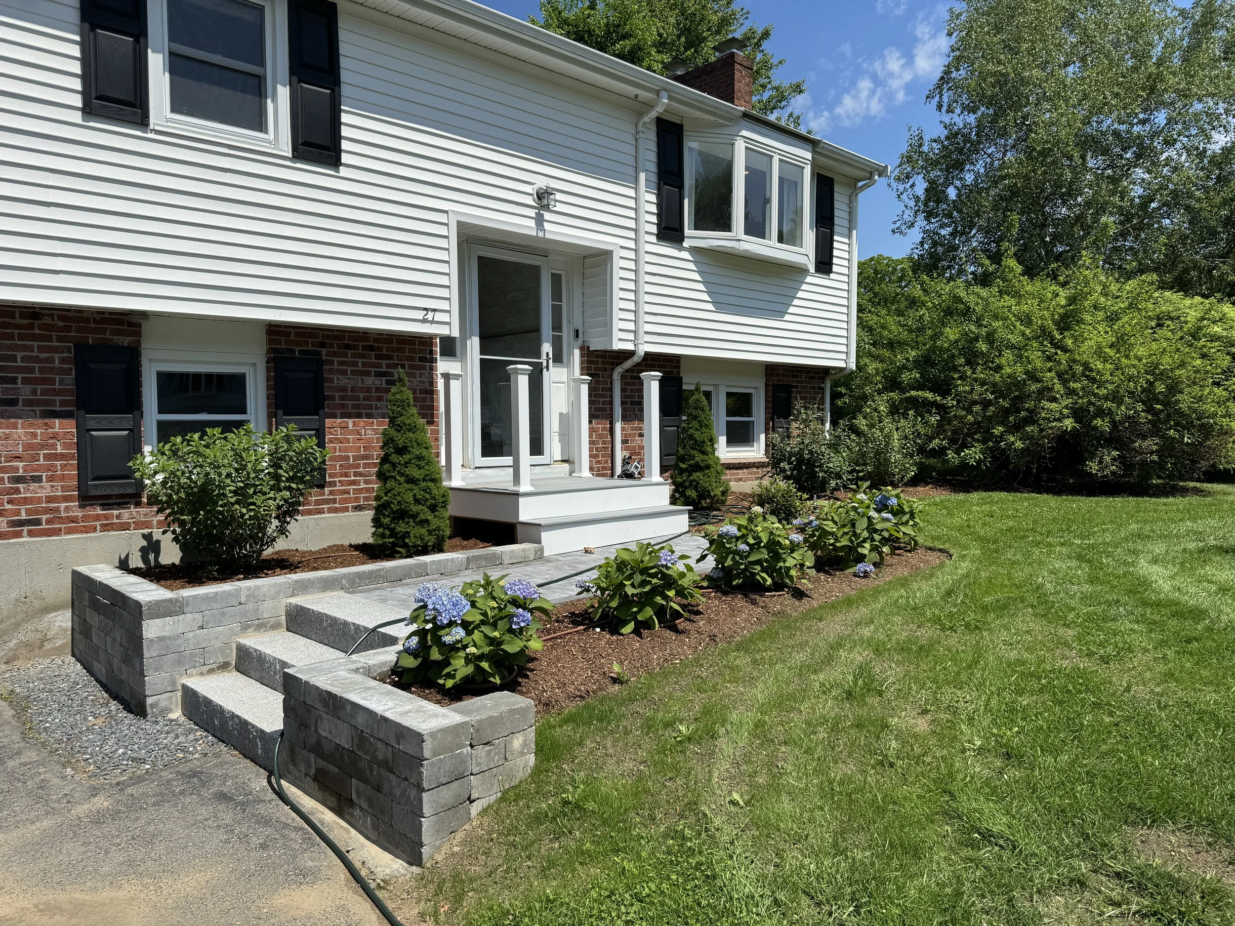 A two-story house with white vinyl siding and red brick foundation, with a small front porch, green lawn, and landscaped garden with blue hydrangeas and shrubs.