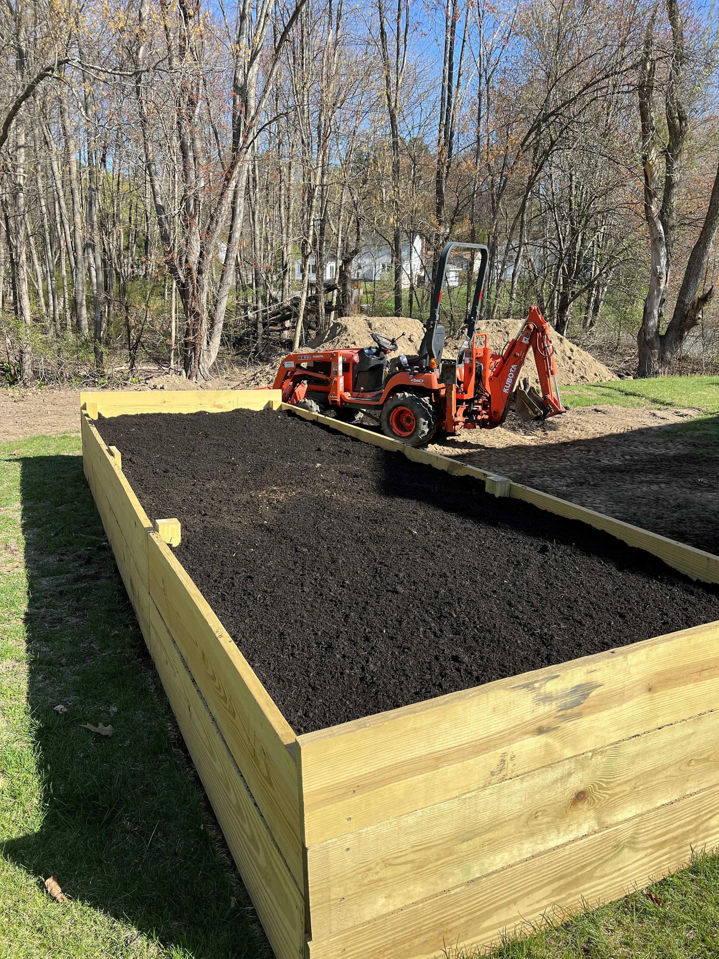 A small orange tractor with a backhoe attachment working in a backyard with a raised garden bed made of wood and freshly filled with soil, surrounded by grass and trees.
