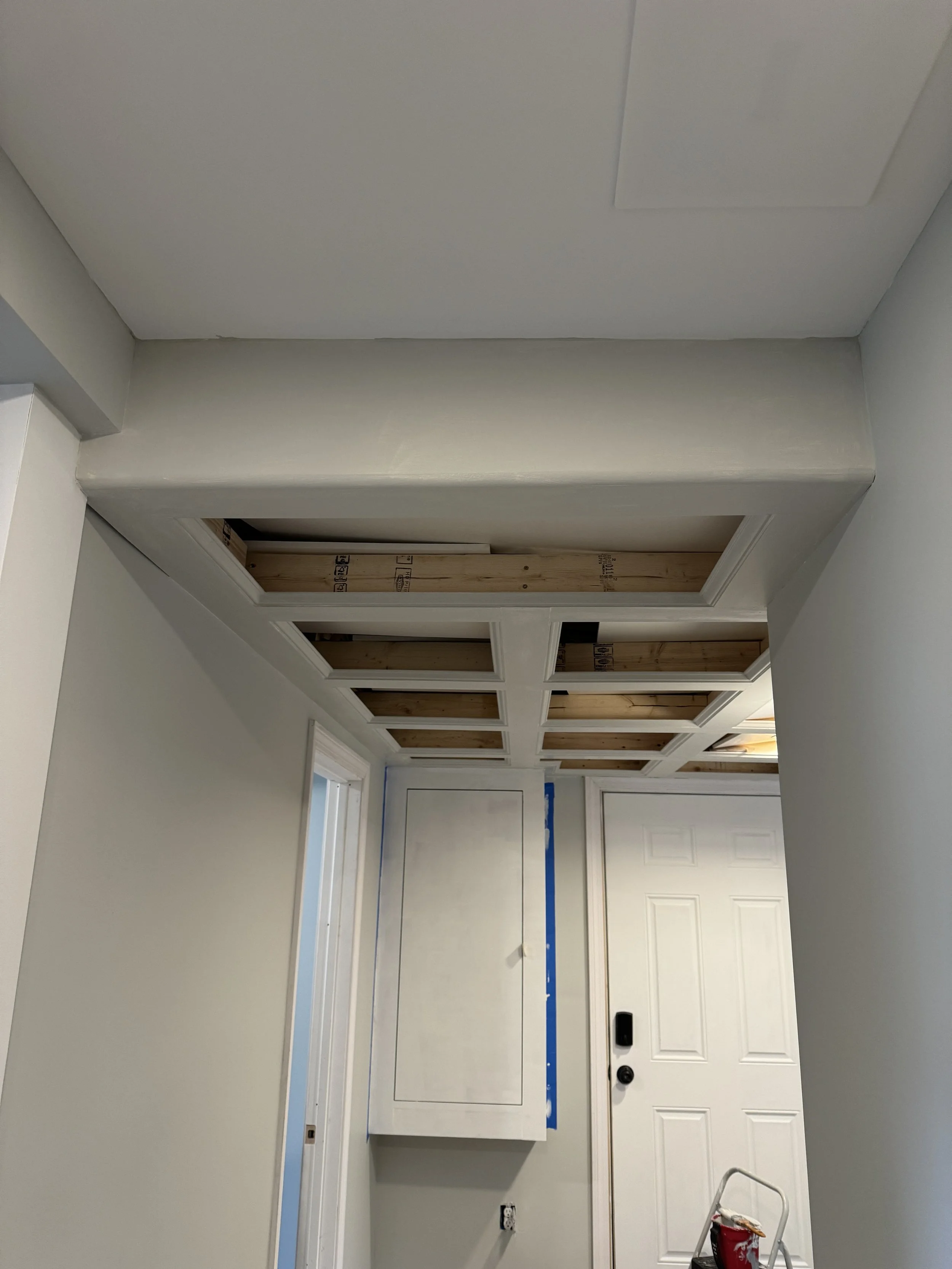 Interior view of a room under construction featuring an unfinished ceiling with wooden framing, a white door, an electrical outlet, and a stepladder with a red bucket on top.