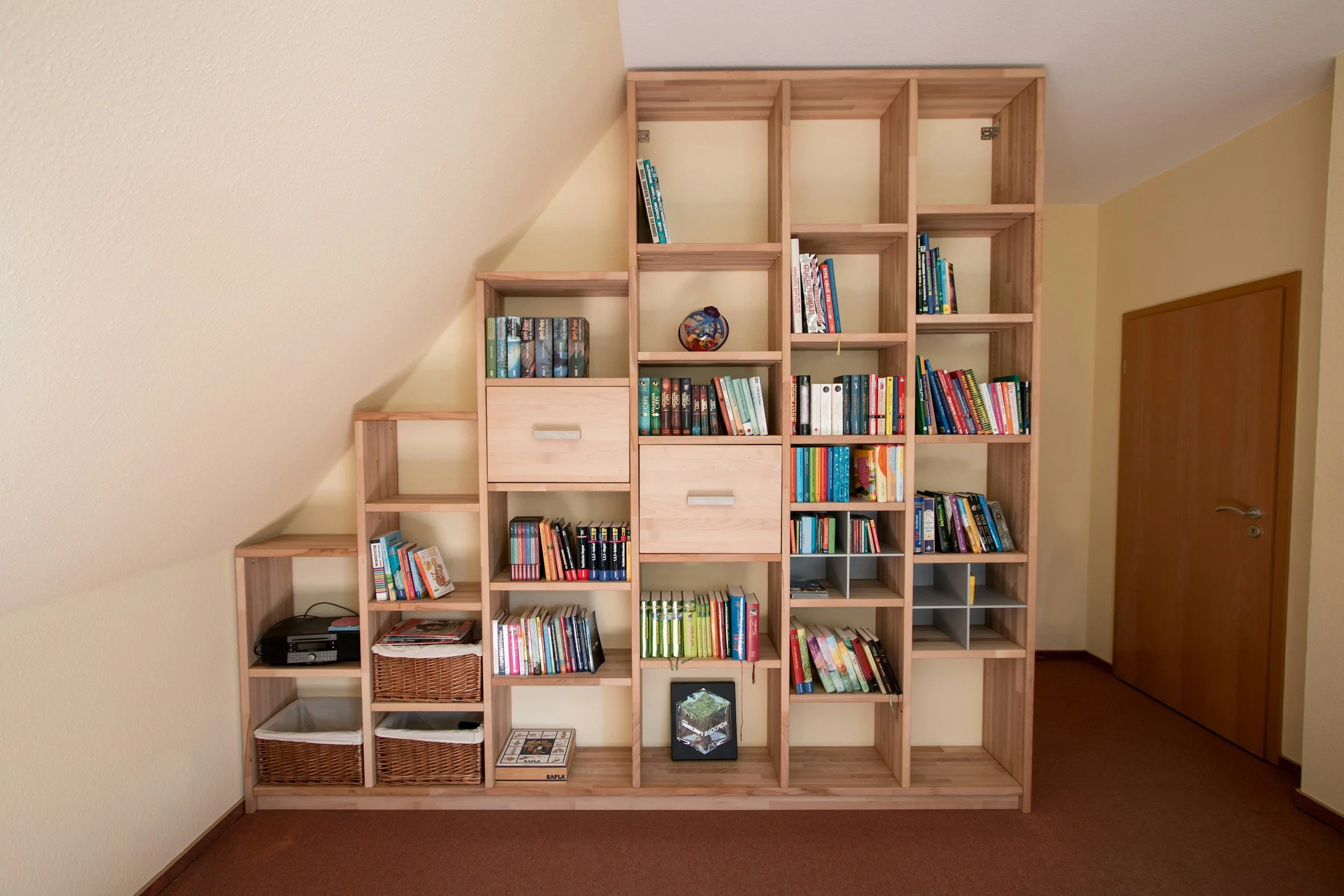 wooden bookshelf with books, drawers, baskets, and decorative items against a beige wall in a room with a brown carpet and wooden door.