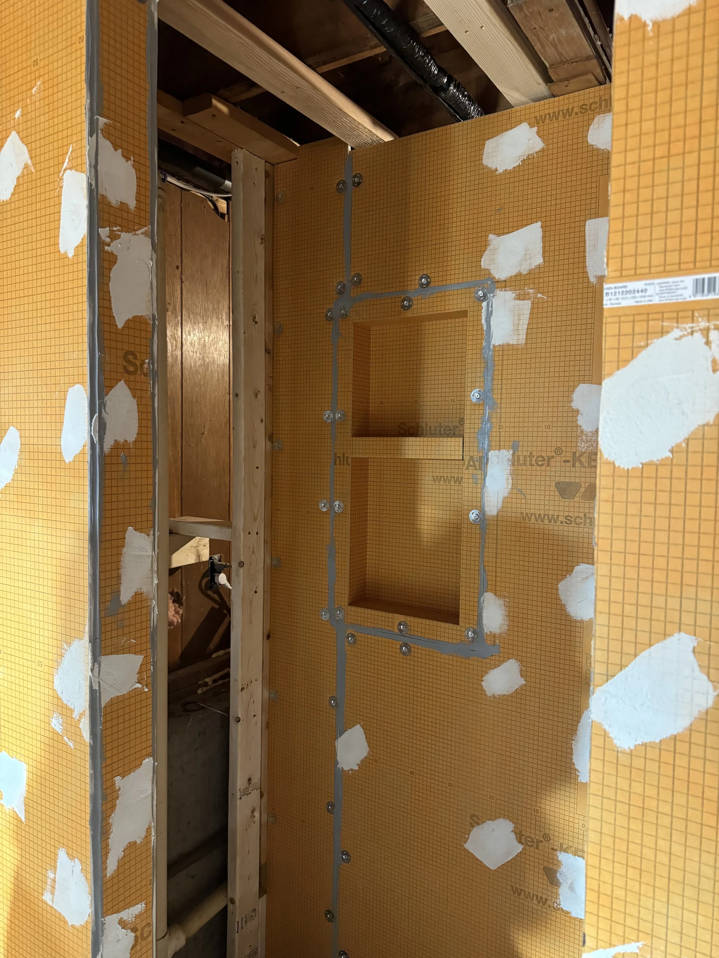 Construction site showing wall with orange waterproofing board, metal electrical conduit, and a rectangular opening for a recessed fixture or access panel, with wooden framing and exposed beams in the background.