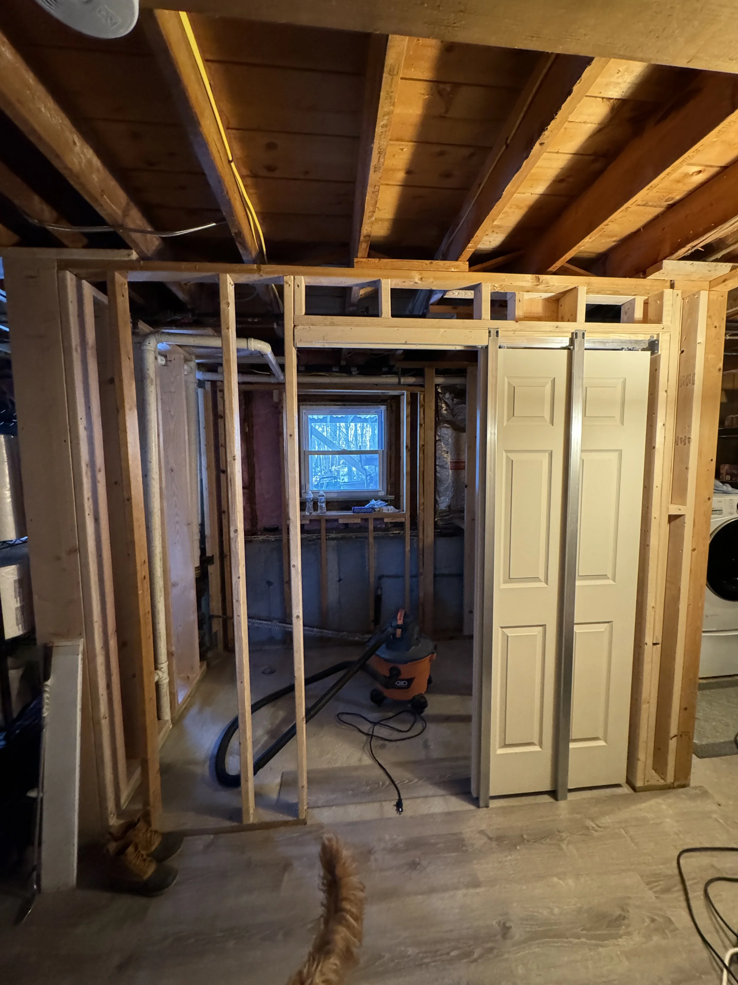 View of a partially framed bathroom under renovation with exposed wooden studs, a small window, and construction tools, with a dog’s tail visible at the bottom.