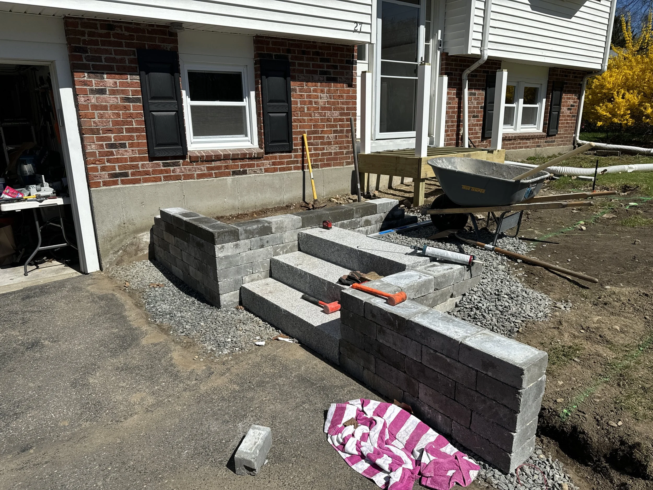 Construction of outdoor steps with gray concrete blocks and masonry tools in front of a brick house with a black shutter window.