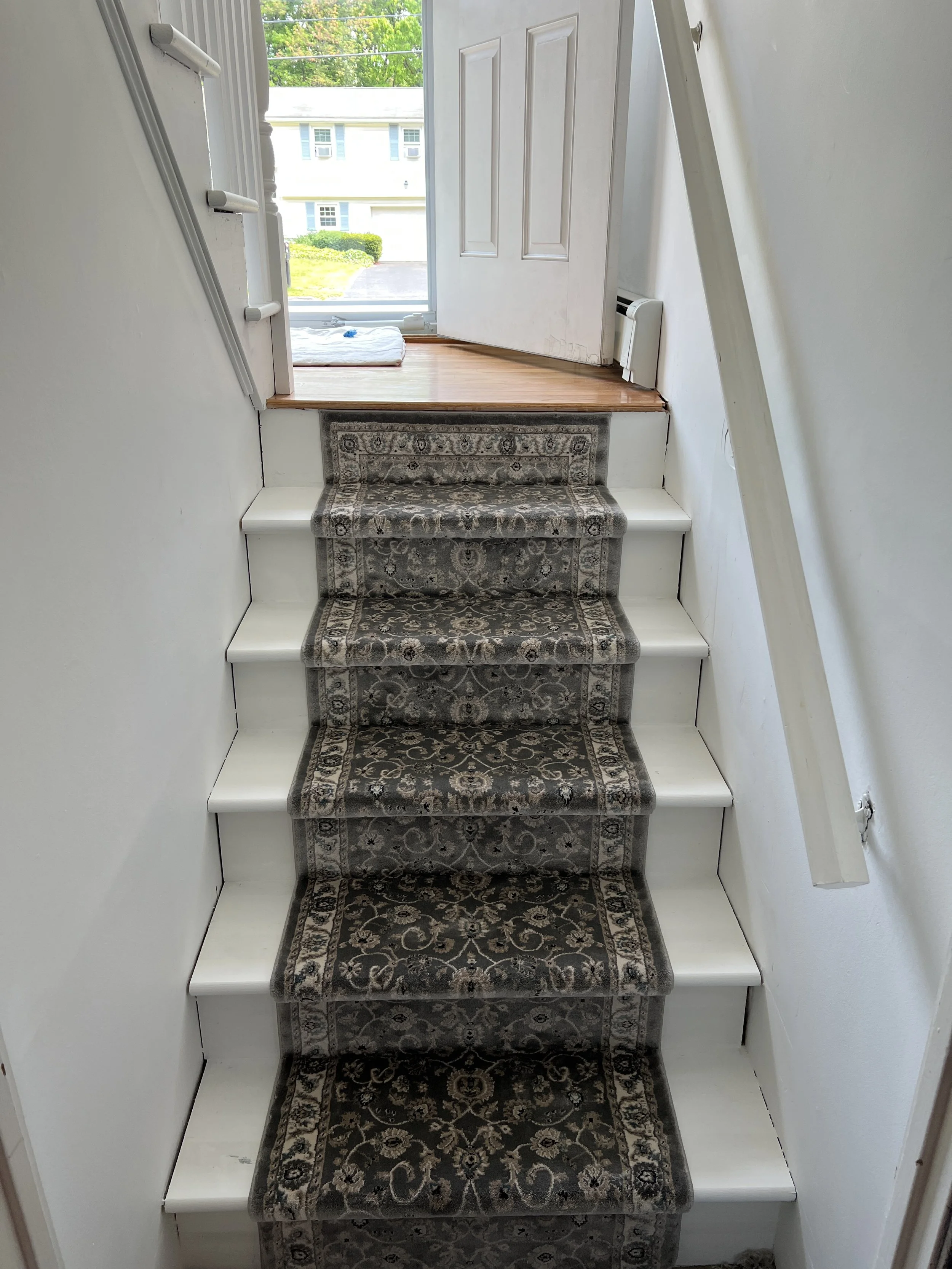 Interior view of a staircase with a patterned carpet runner, leading to a small area near a door that opens to a porch with a view of neighboring houses.