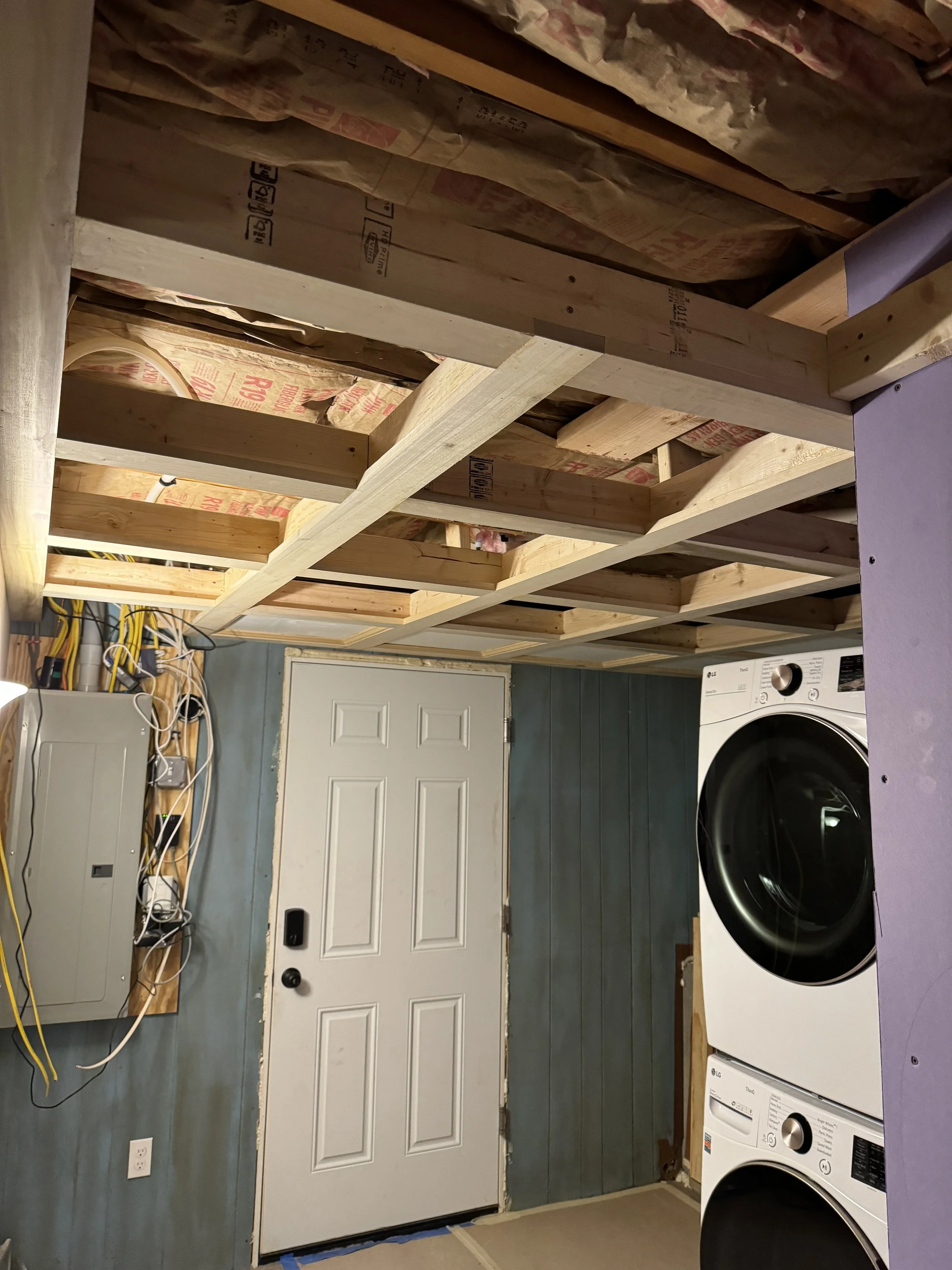 Laundry room under construction with exposed wooden framing on ceiling, a white door, a washing machine and dryer, and an electrical panel with wires.