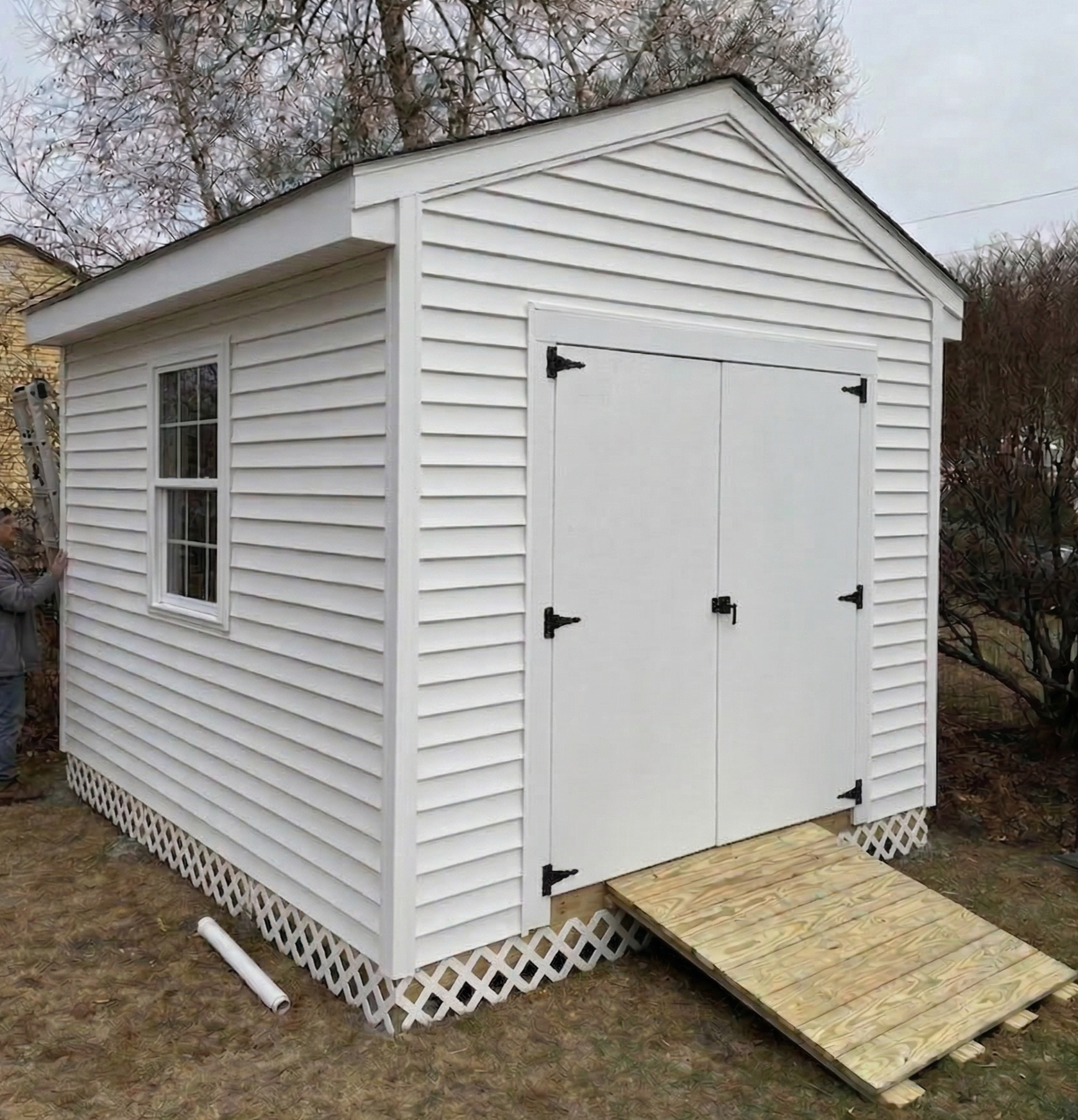 A person installing a small white shed with vinyl siding, two window panels, and black hinges, on a wooden platform in a yard with trees and a lattice skirting at the bottom.
