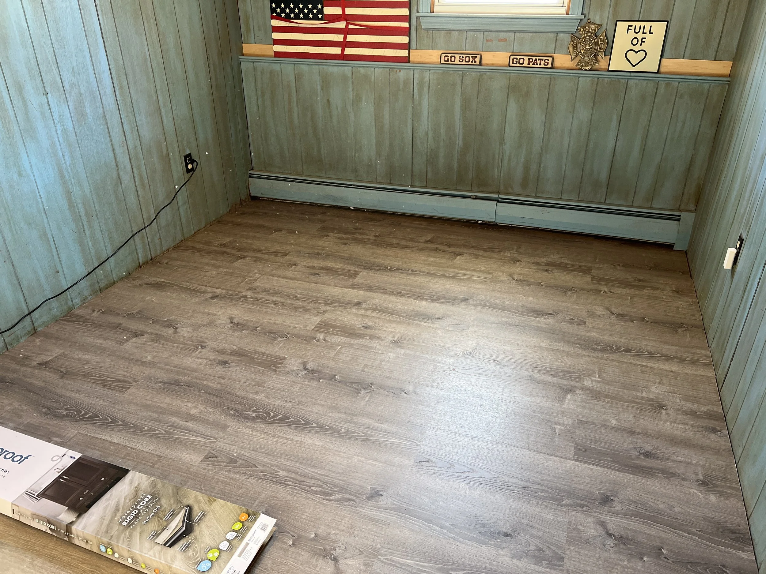 Empty room with wood-look flooring, green wooden walls, a window with patriotic decorations, and a box of flooring on the floor.