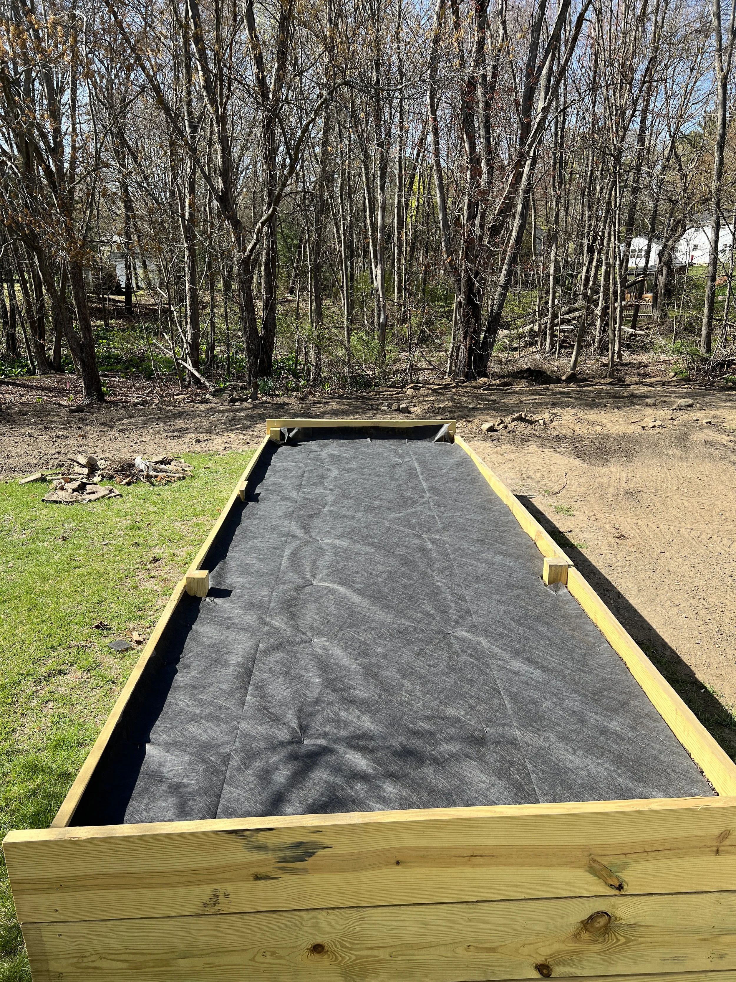 A rectangular wooden frame with a black liner inside, set outdoors on grass and dirt, with trees in the background.