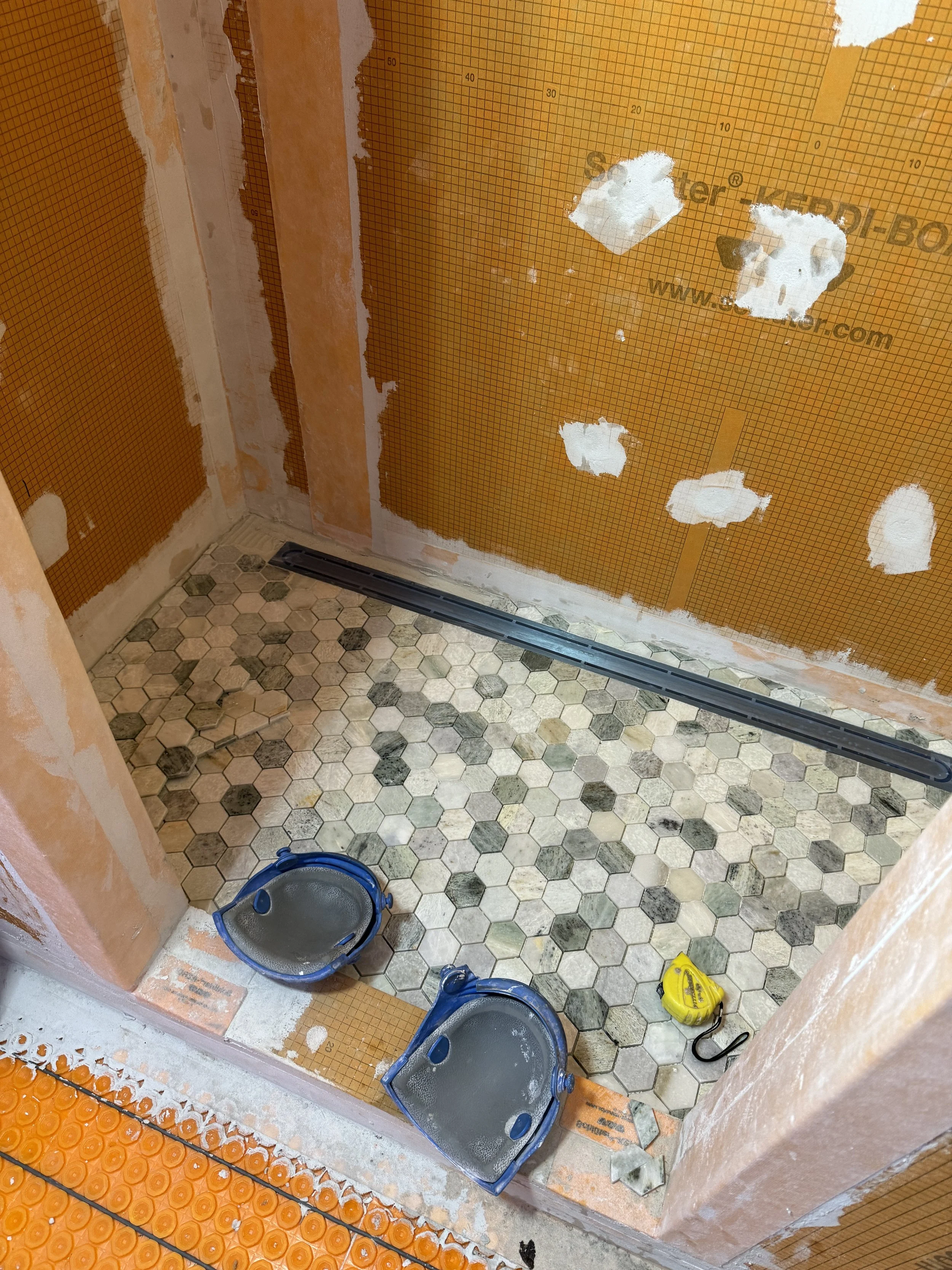 A bathroom under construction with unfinished walls, hexagon tile floor, two blue wall-mounted toilet bowls, and a yellow measuring tape on the floor.