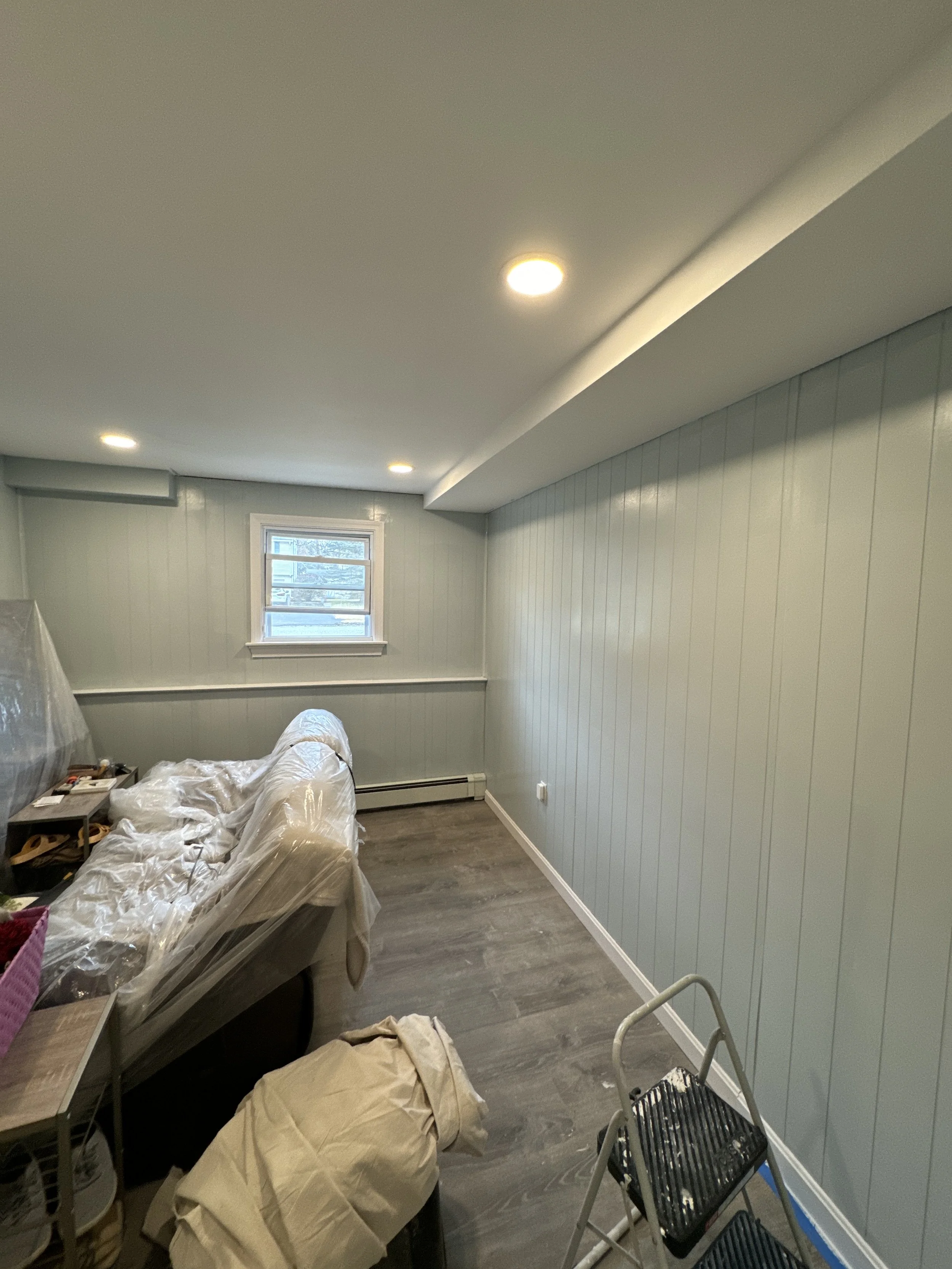 A small room with light blue paneled walls, a small window, and a ceiling with recessed lighting. There is a plastic-covered couch, a step ladder, and some miscellaneous items.