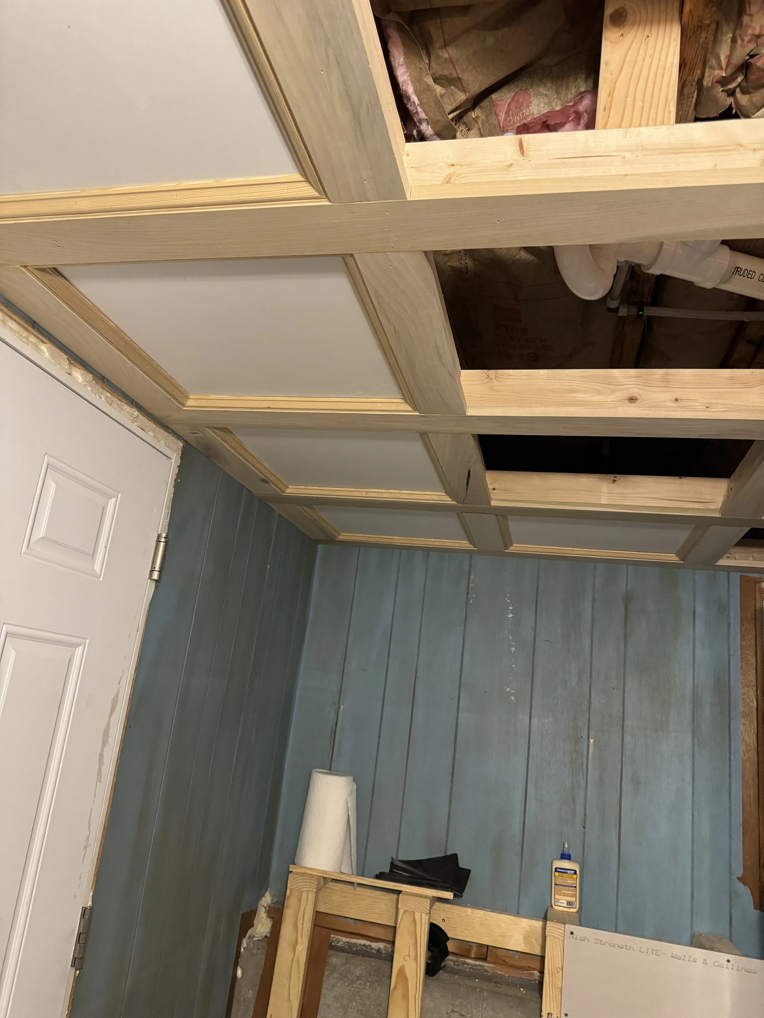 Interior room under construction with wooden framing and blue paneled walls, a white door, and construction materials on the floor.