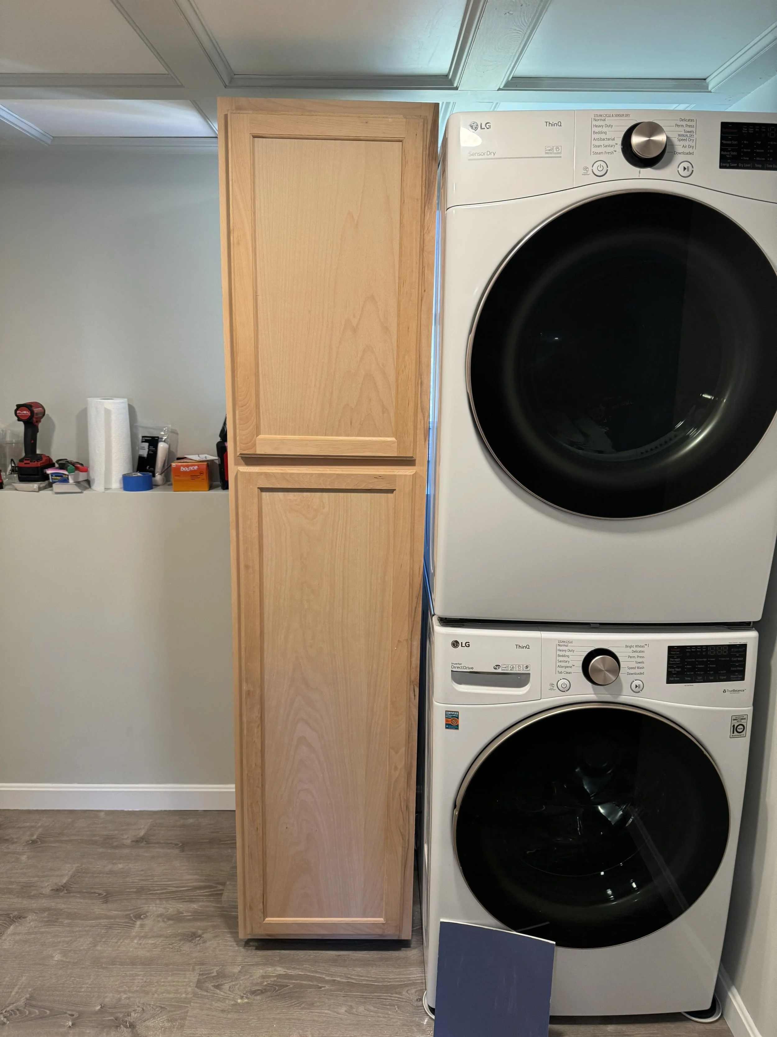 A laundry room with a stacked LG washer and dryer on the right side, a wooden cabinet in between, and a cluttered shelf on the left with tools, paper towels, and other items.
