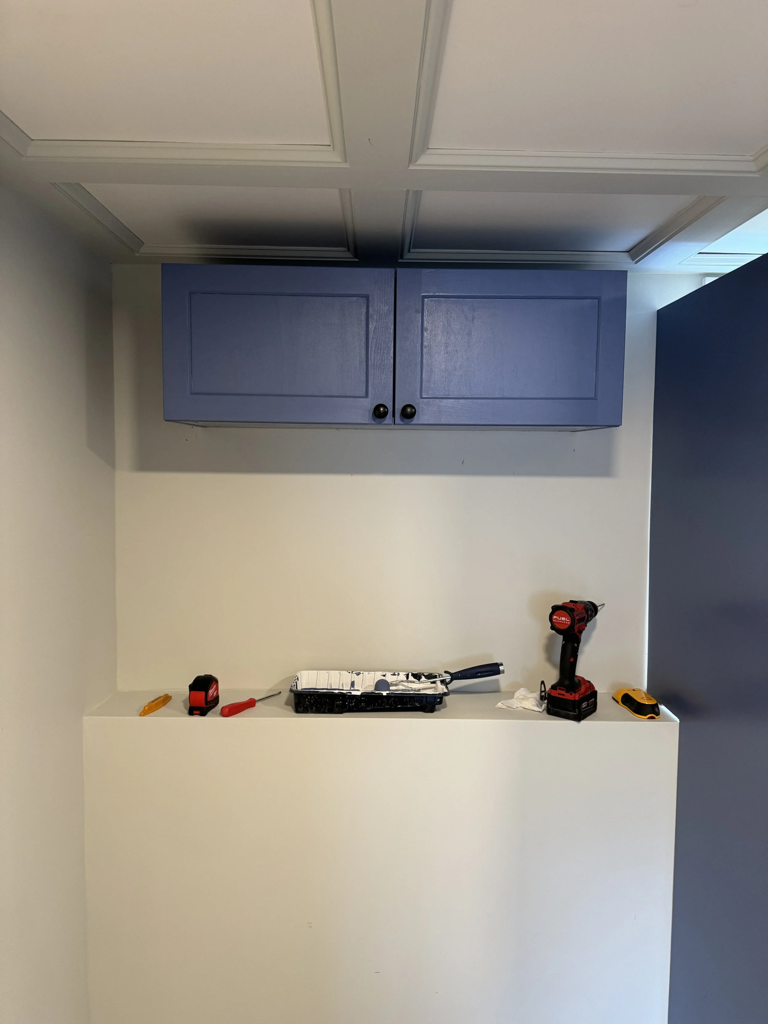 Empty home improvement workspace with tools including a tape measure, screwdriver, drill, paint roller, and tape measure on a white ledge, with a blue cabinet above.