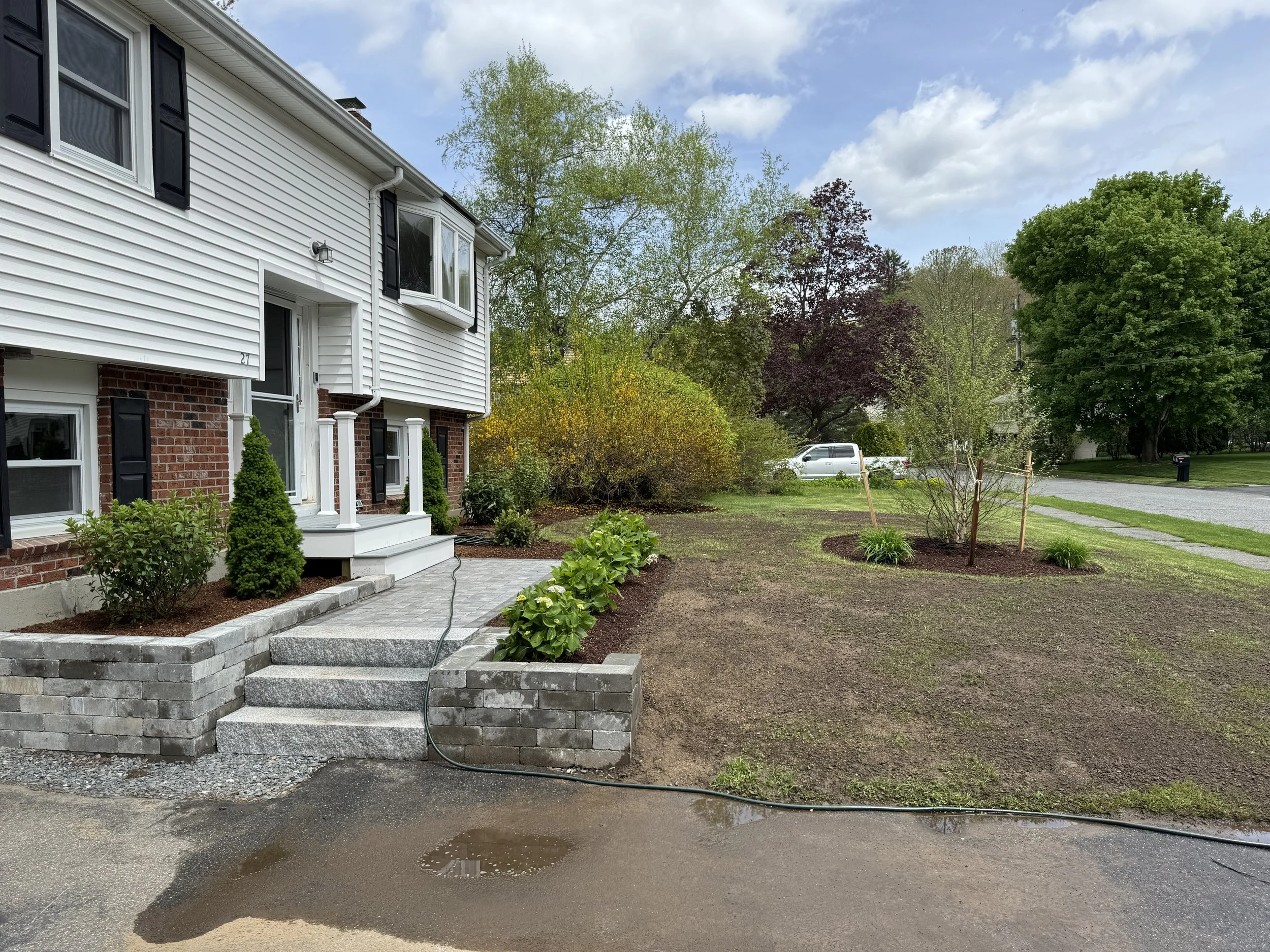 A house with white siding and brick foundation, with a garden bed with small bushes and a small tree, and a dirt yard area, some within new mulch. There is a small stone patio with steps leading to the porch area, and a hose on the ground. In the bac