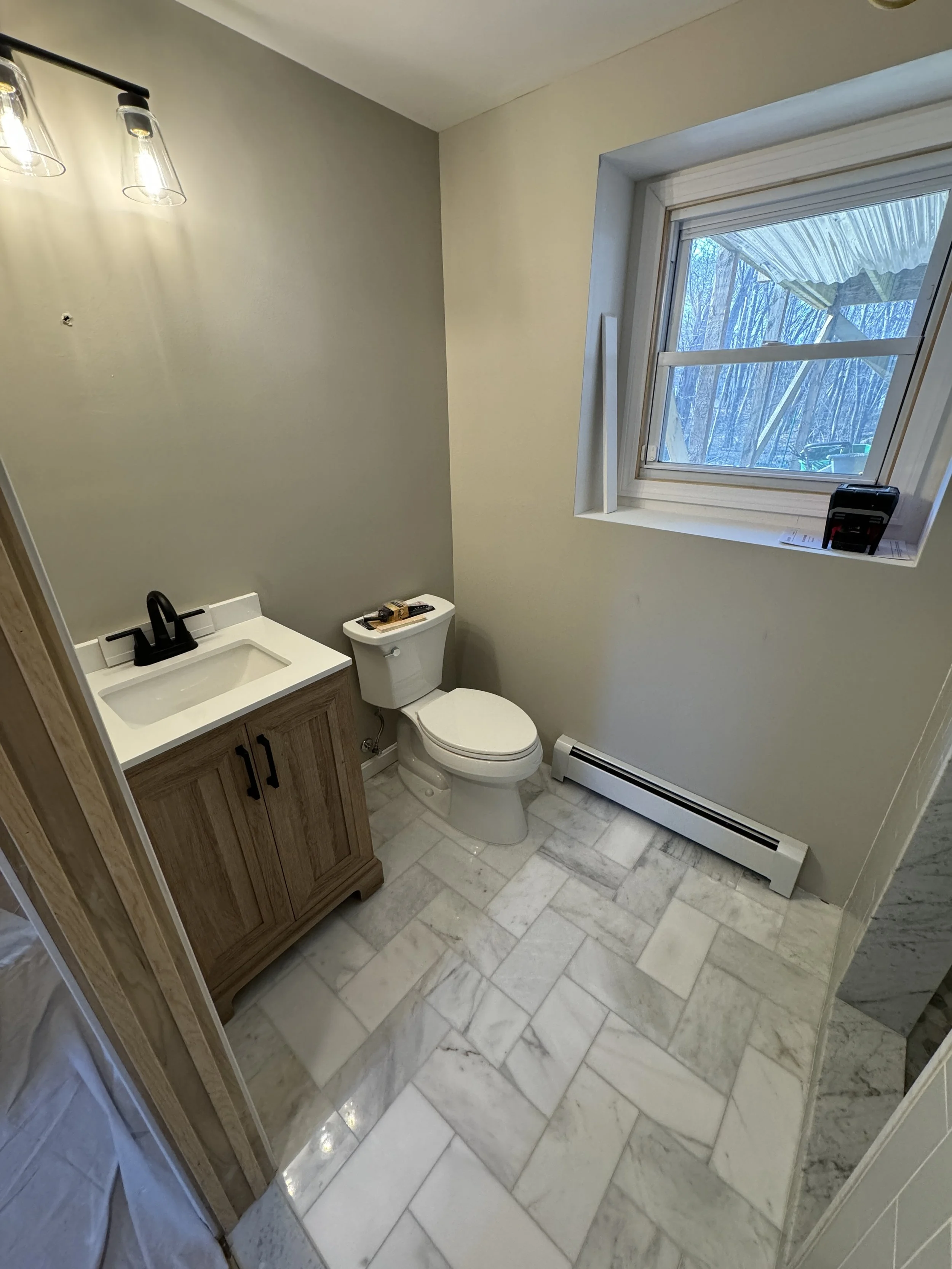 Small bathroom with marble tiled floor, a wooden vanity with a black faucet, a white toilet, and a window with an outdoor view. There is a light fixture with three bulbs on the ceiling.