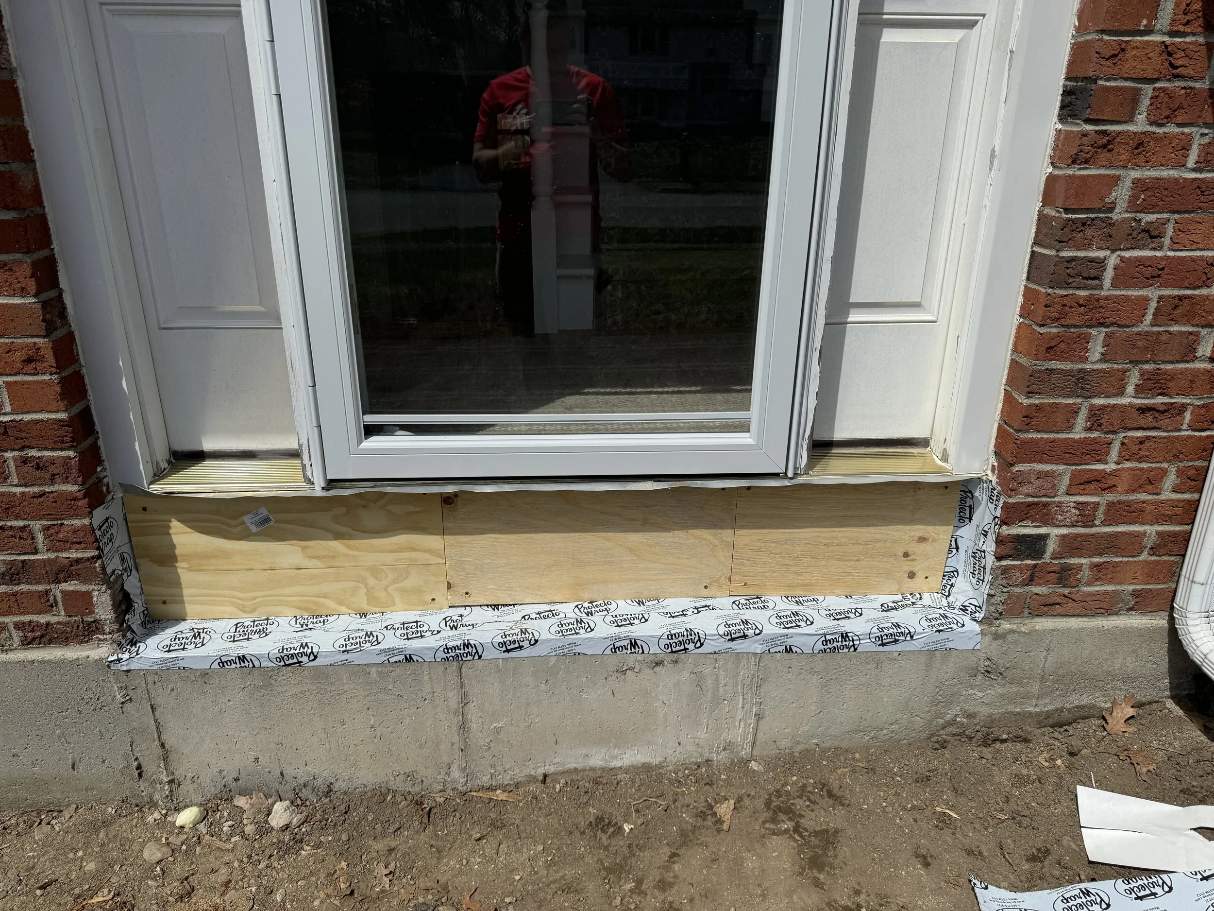 Front door with a new lower frame under construction, surrounded by brick wall and house siding, with construction materials and dirt on the ground.