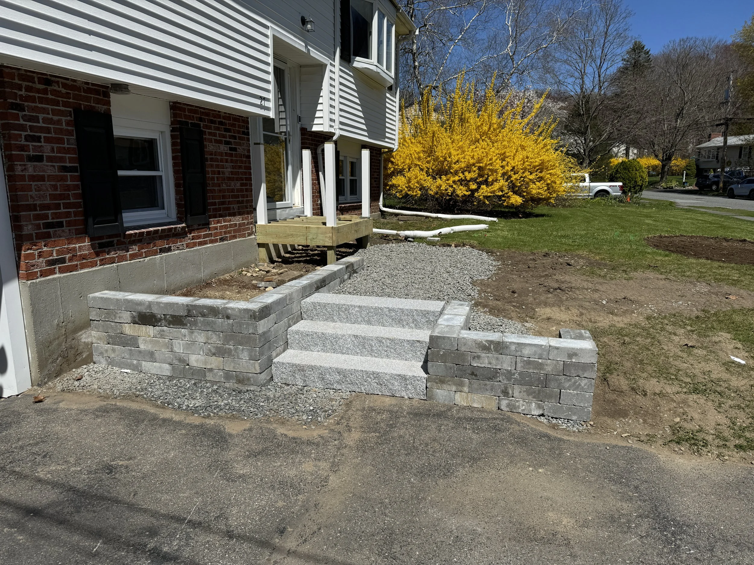 Newly constructed front steps with stone slabs and brick sides leading to a house with white siding and black shutters, with a landscaped garden to the side featuring yellow flowering shrubs and trees.