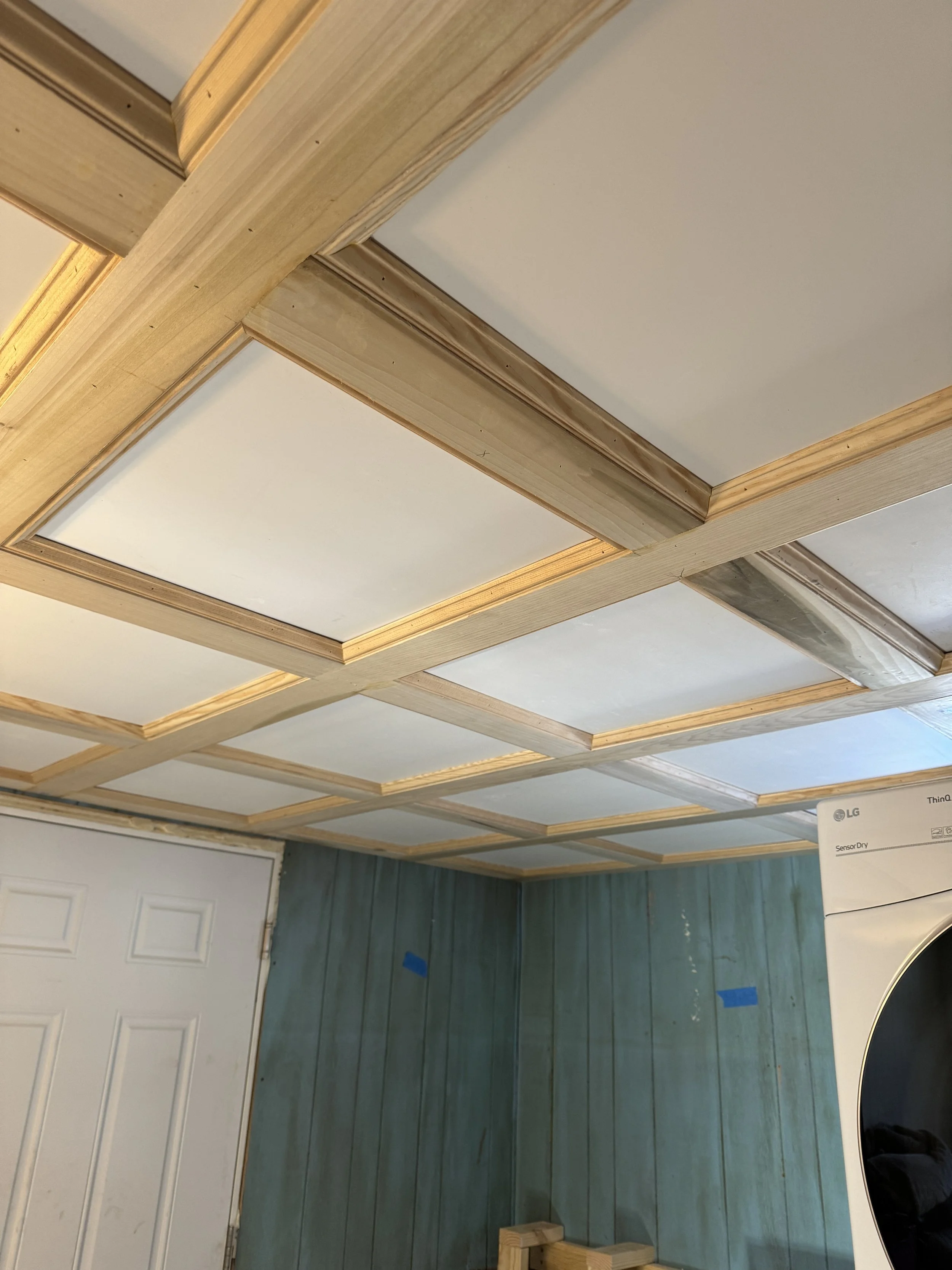 Ceiling with wooden grid framework and white panels in a laundry room with light blue shiplap walls and a white door.