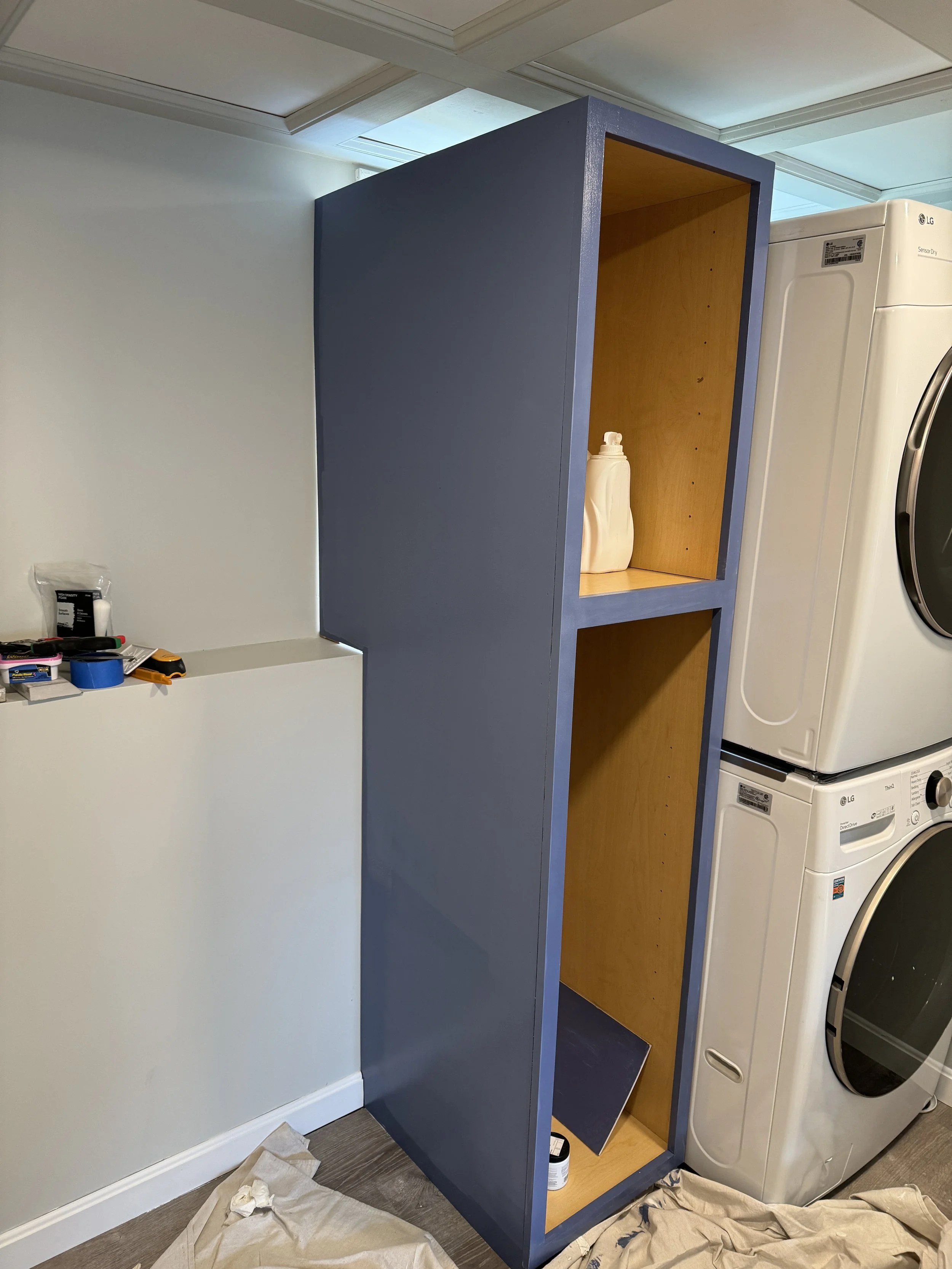 A partially painted tall blue wooden shelving unit in a laundry room, with two shelves and a visible bottle of detergent on the upper shelf. Laundry appliances are on the right, and a white wall with tools and supplies is on the left.