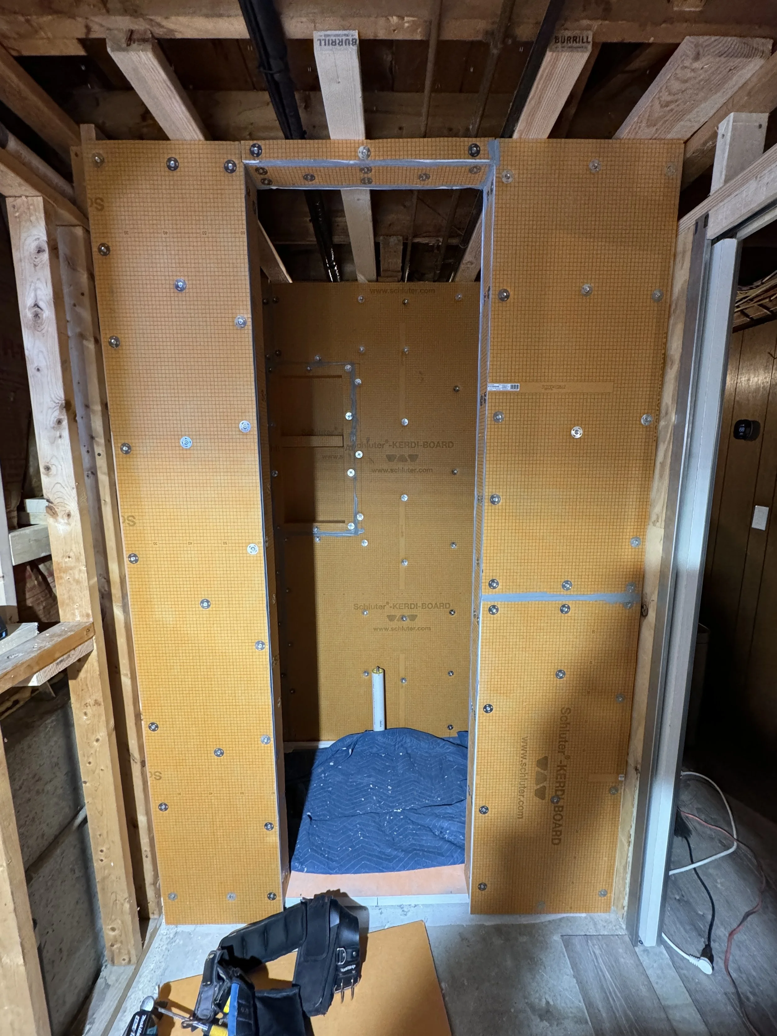 Framing of a small bathroom or closet under construction, with yellow KERDI-BOARD panels attached to the wall and ceiling, electrical conduits, and a plastic sheet on the floor.
