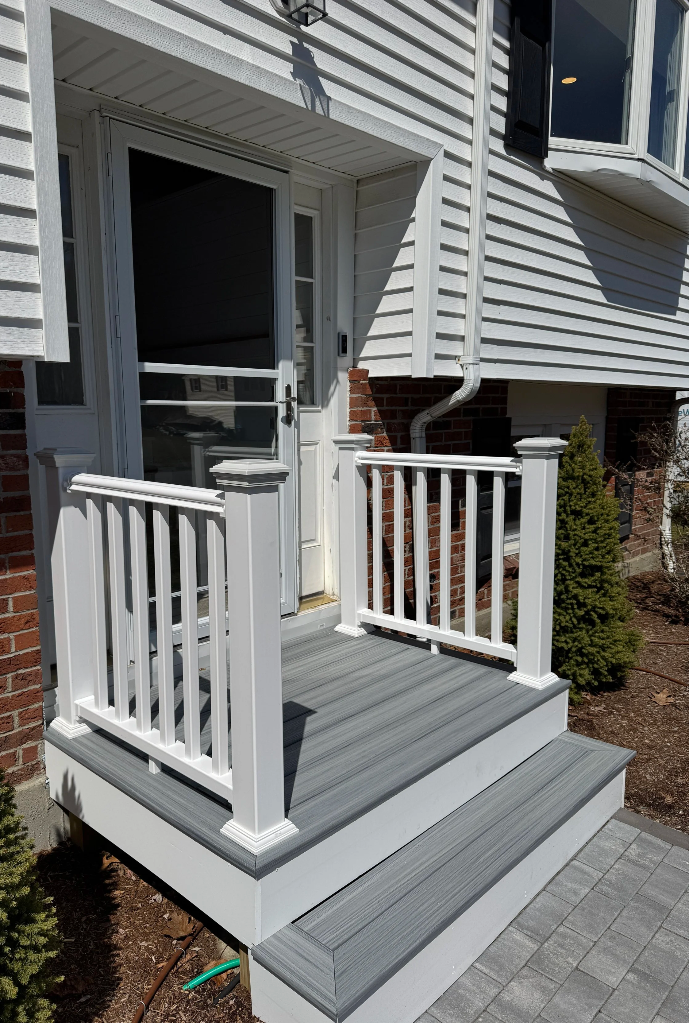 Small front porch with gray decking, white railing, and steps leading to a concrete walkway, attached to a house with white siding and brick base.