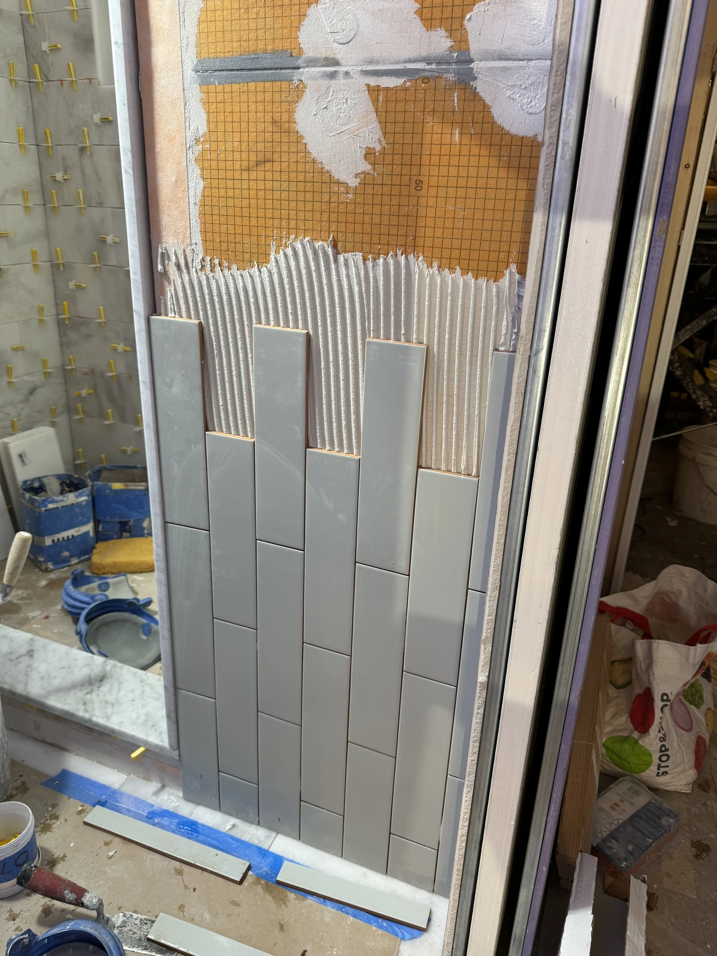 Partial wall with white subway tiles being installed, with some adhesive and lath visible behind the tiles, and construction tools and supplies scattered around.