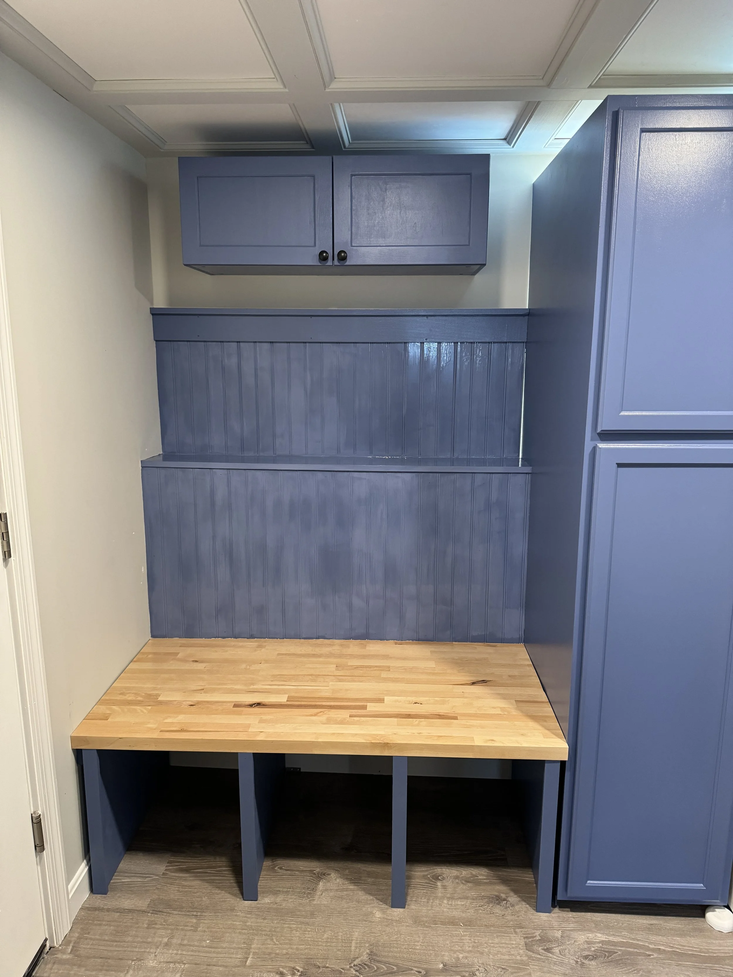 Blue built-in cabinetry with upper and lower storage compartments, a wooden bench seat with open storage underneath, and a beadboard backing on the wall.