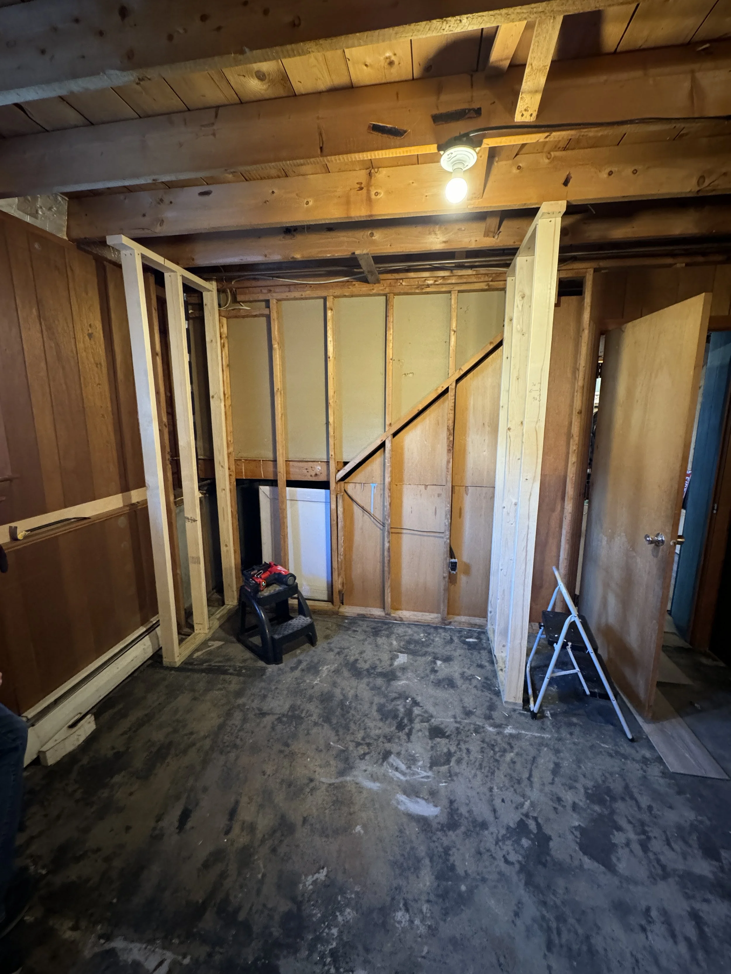 Interior view of a room under construction with exposed wooden framing, a partially finished wall, a small black step stool, and a white folding step ladder on a dirty concrete floor.