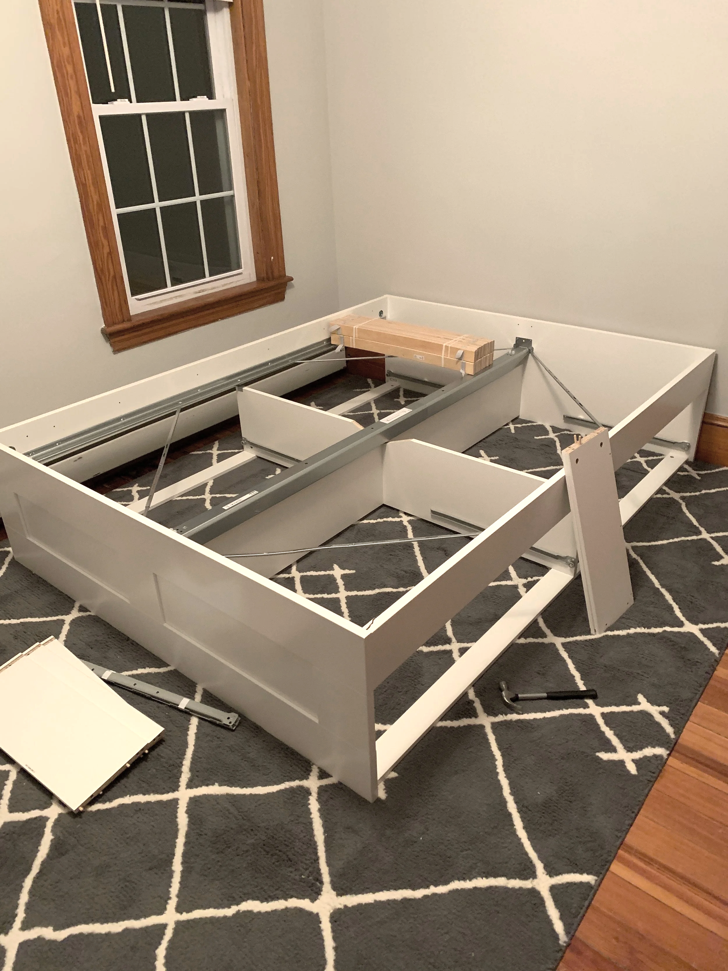 Room with a disassembled white bed frame on a patterned gray and white rug, with a hammer on the floor and a window with wooden trim.