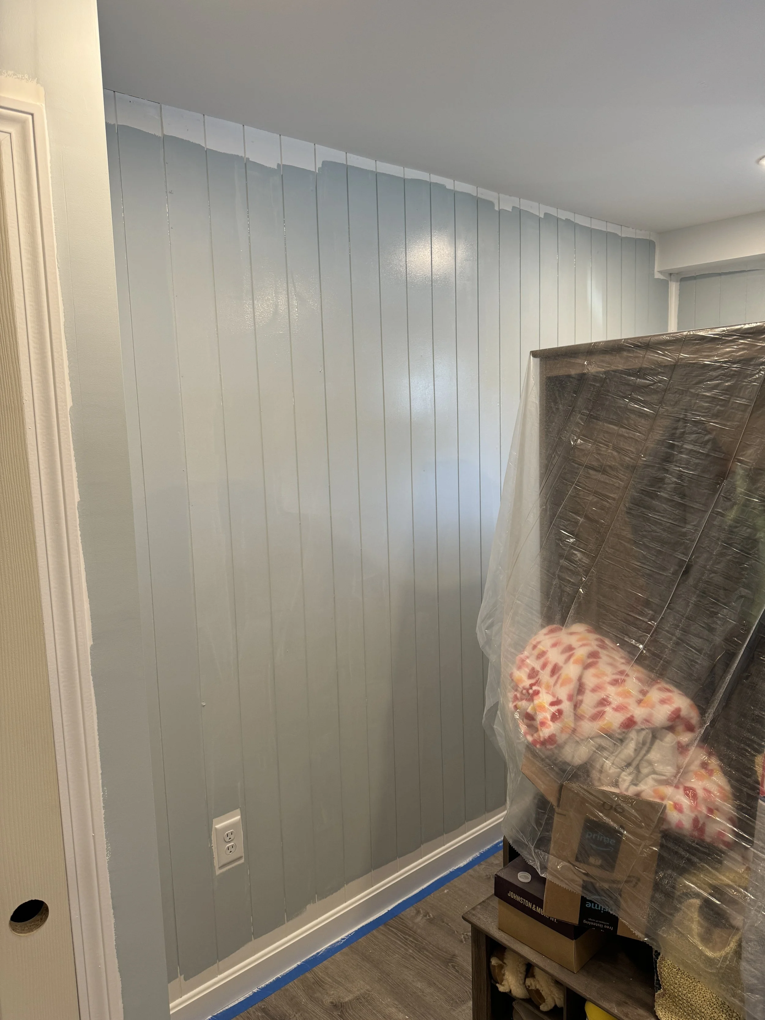 Interior of a room with a partially painted light blue wall, a white electrical outlet, and a piece of furniture covered in plastic with a blanket on top.