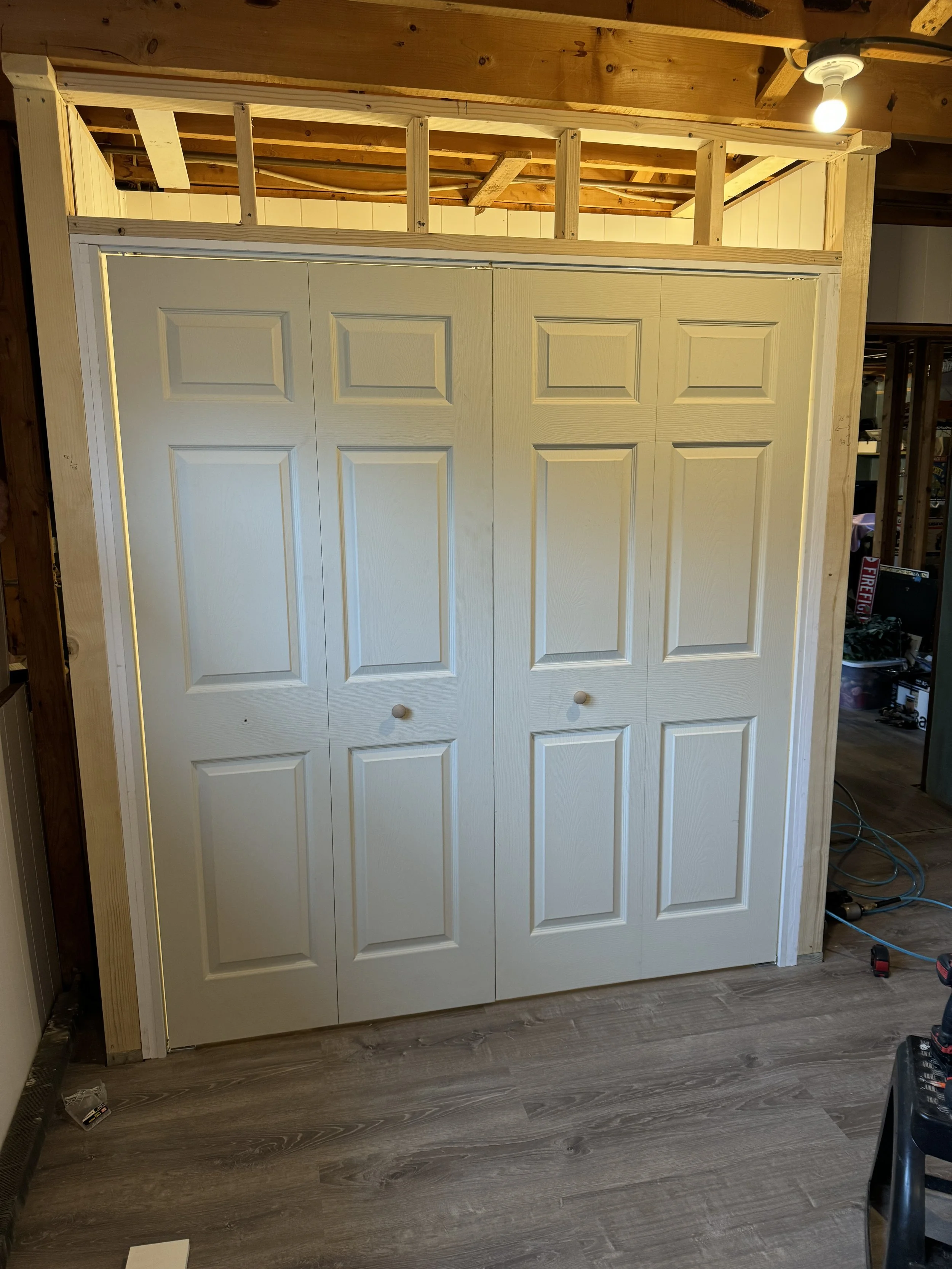 A double closet with white paneled doors in a room under construction, with wooden framing and an unfinished ceiling.