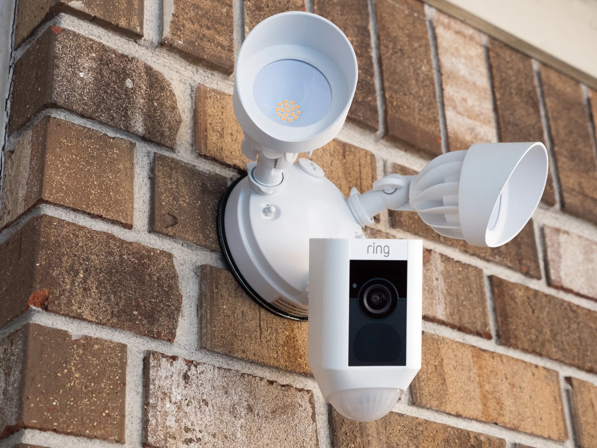 Security camera system mounted on a brick wall, consisting of two white camera units and a sensor or a light attached to a white fixture.