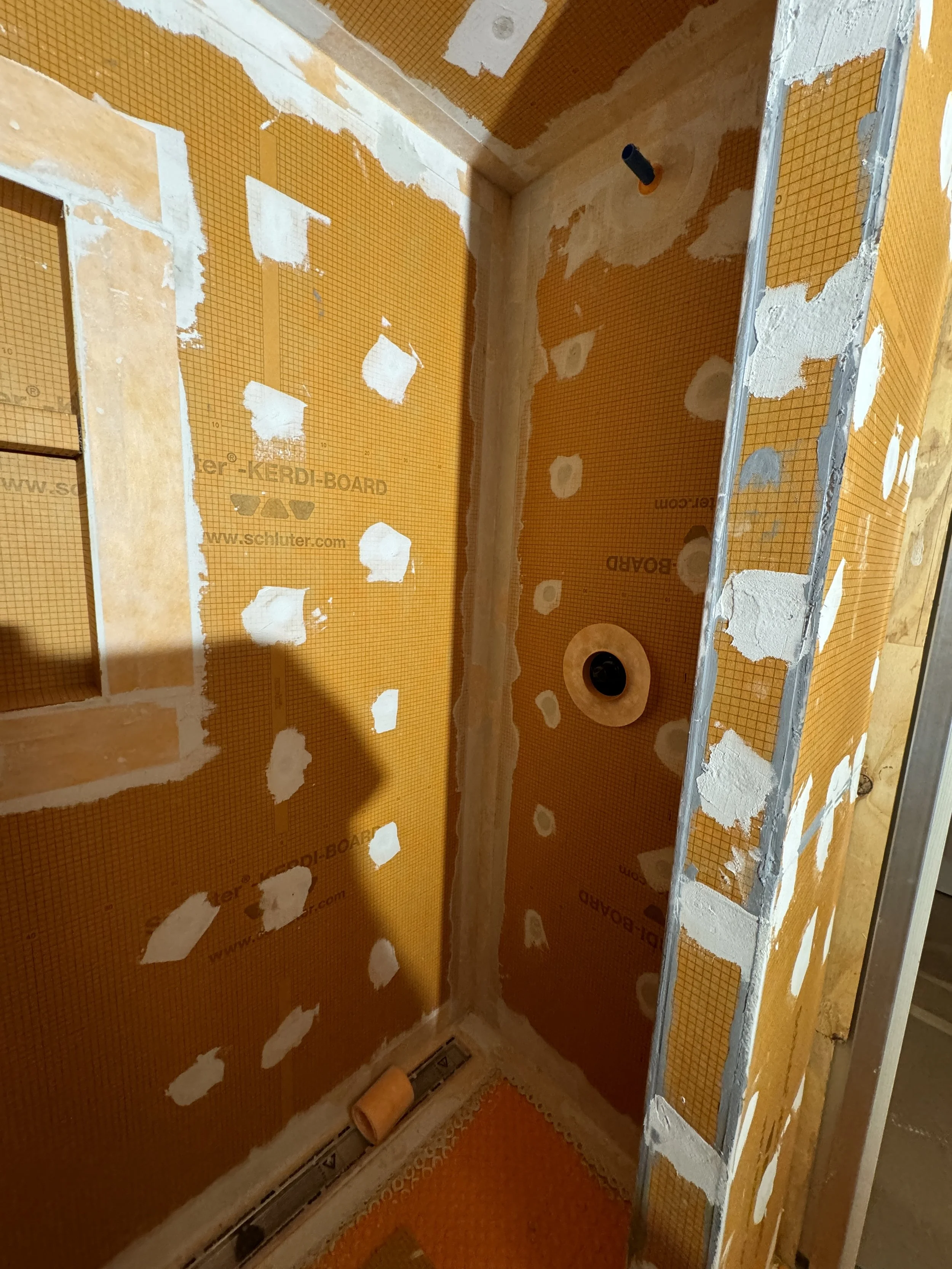 Bathroom under construction with orange waterproof wallboard, patches of drywall mud, a pipe in the wall, a circular drain or vent, and an orange floor with a vent.