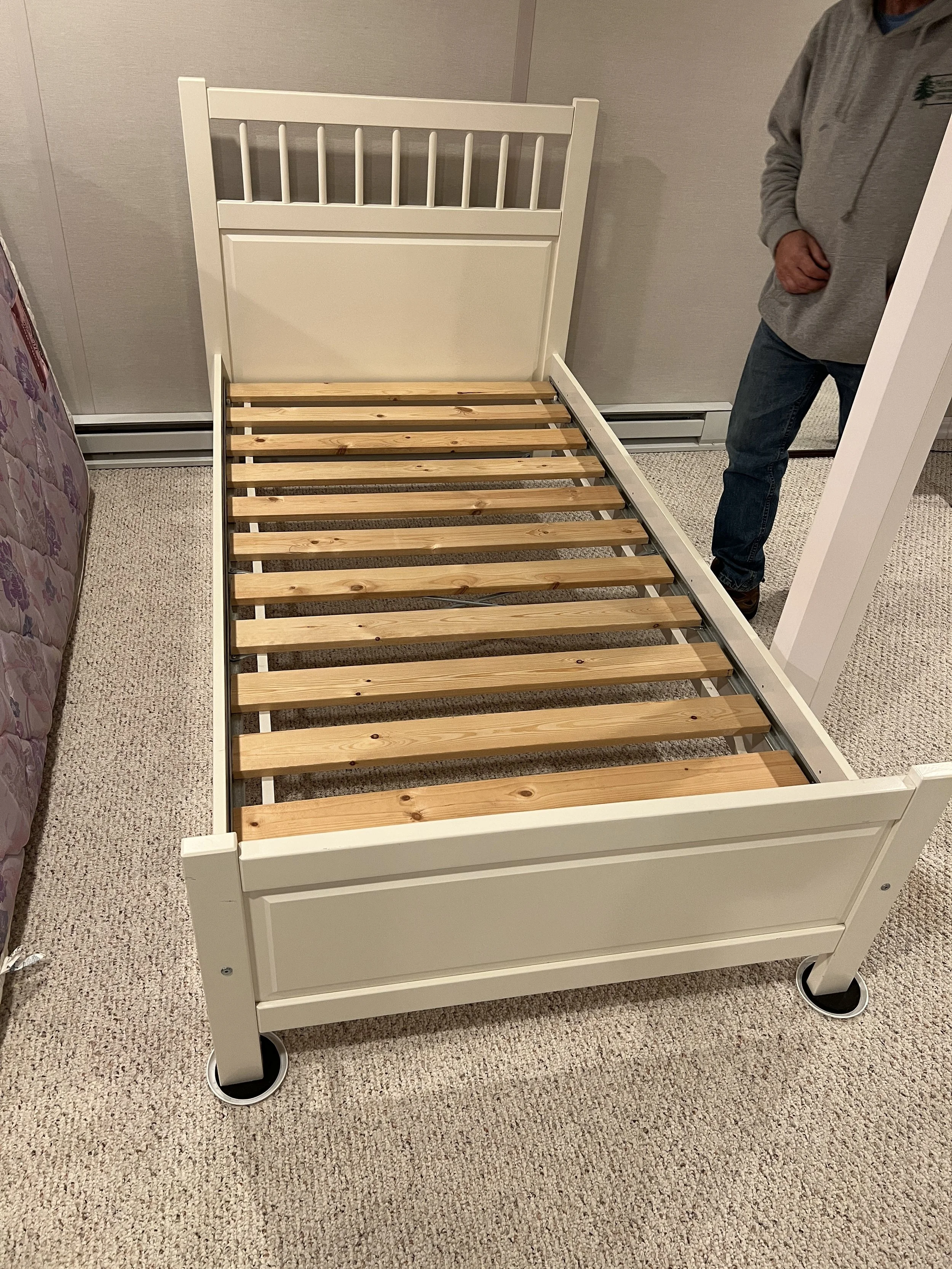 Unassembled white bed frame with wooden slats on a beige carpeted floor in a room, with a person partially visible on the right side.