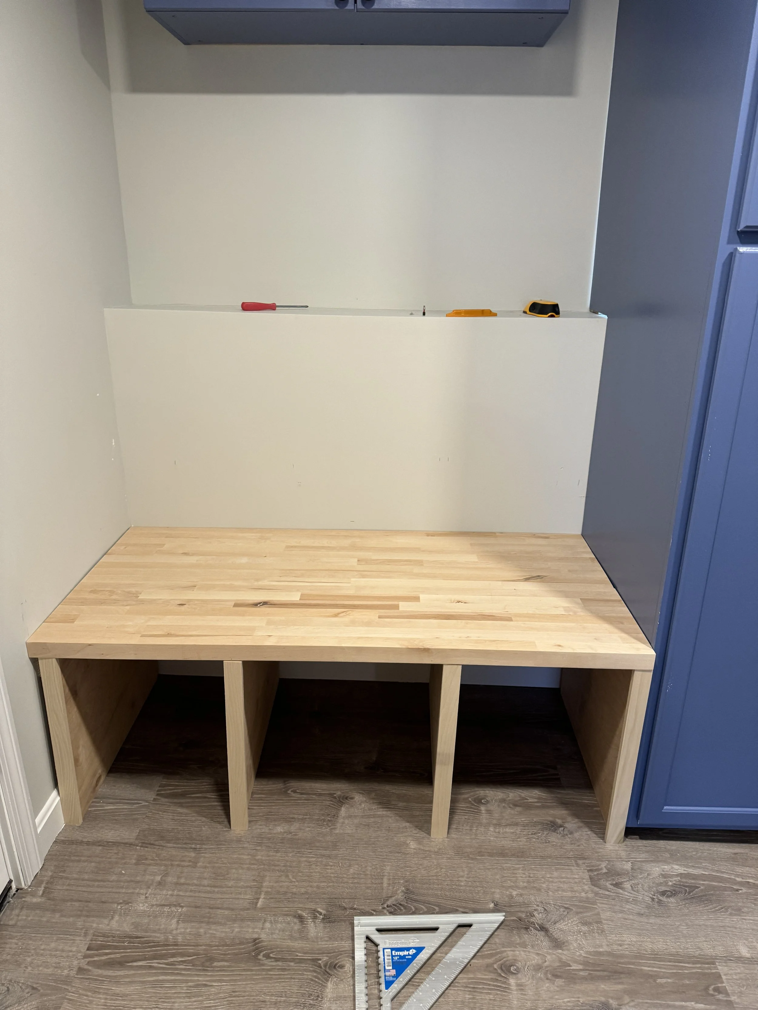 Wooden bench with open cubbies underneath, placed against a wall in a room under construction. Tools are on a white shelf above the bench, and a blue cabinet is on the right side of the image. A carpenter's square and level are on the floor in front 