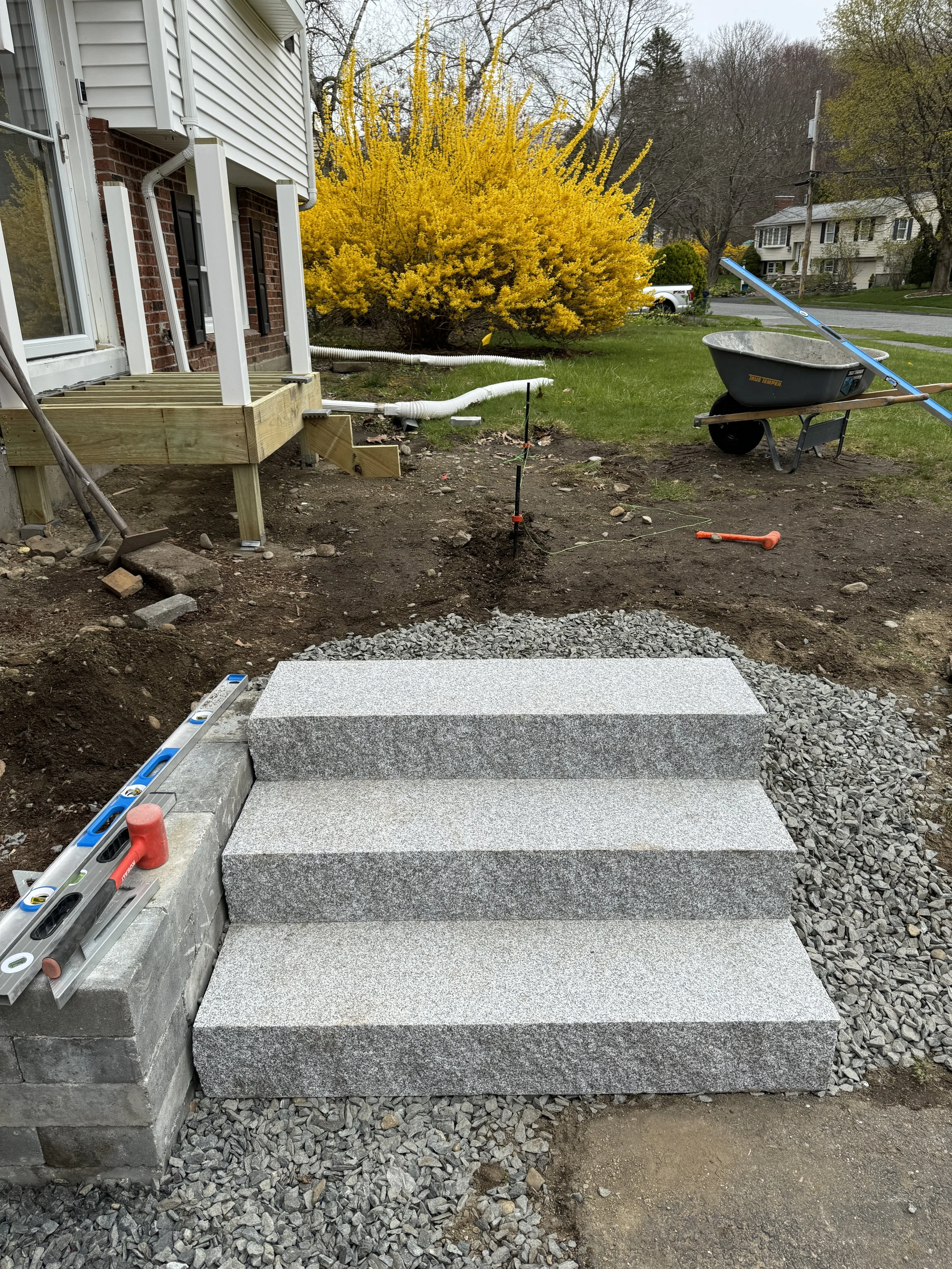 Newly constructed stone steps in front of a house with construction tools around, including a level, rubber mallet, and wheelbarrow, with a yellow flowering bush and trees in the background.