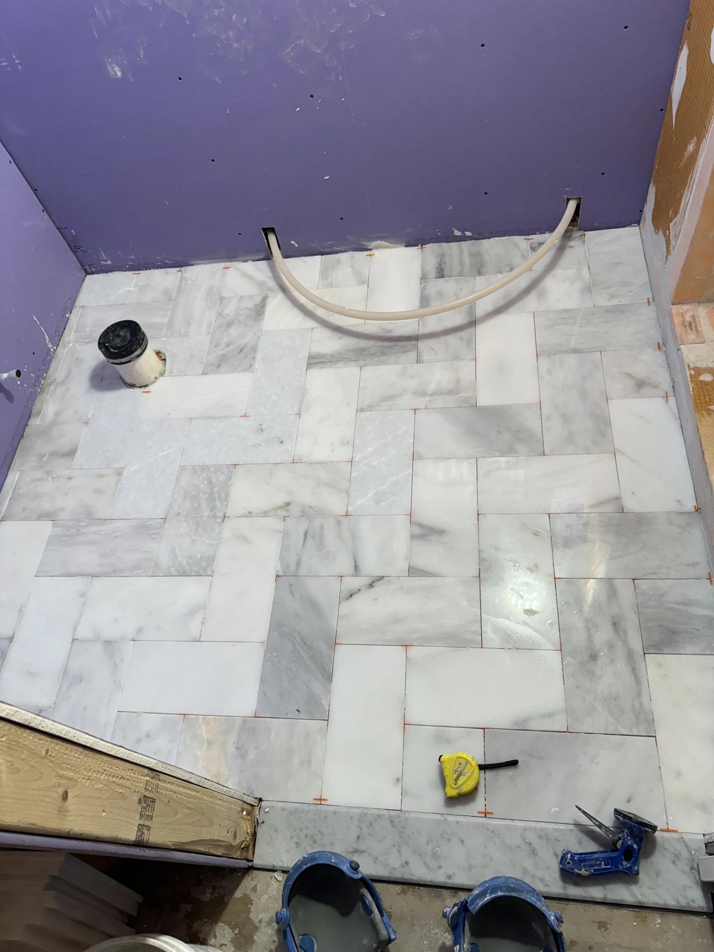 Marble tile flooring installation in progress with tools, plumbing pipe, purple wall, and unfinished construction area.