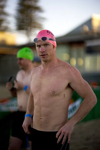A shirtless male swimmer wearing a pink swim cap and goggles, standing outdoors with a focused expression. In the background, another swimmer in a green swim cap is visible, along with a building and trees.
