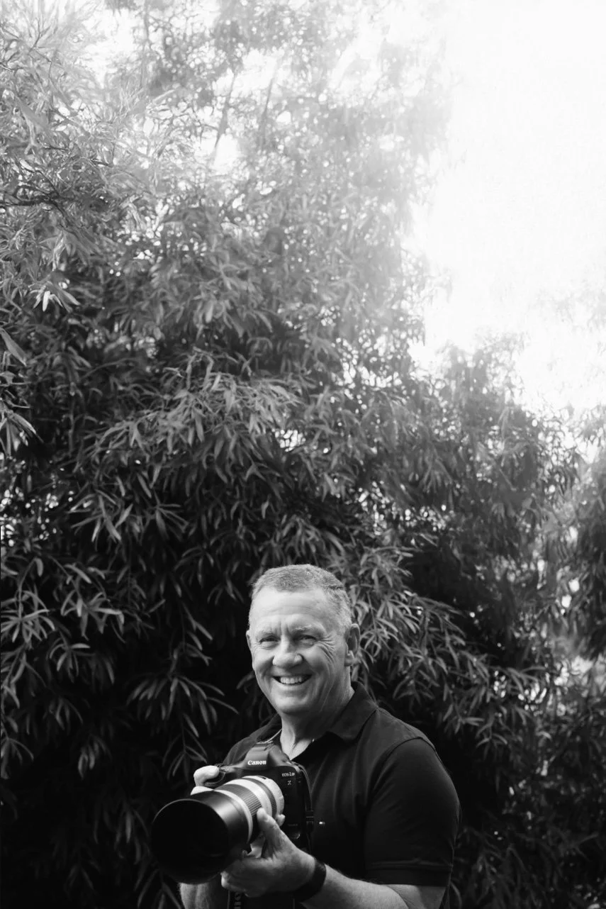 Smiling man holding a camera with a large lens outdoors in front of dense foliage.