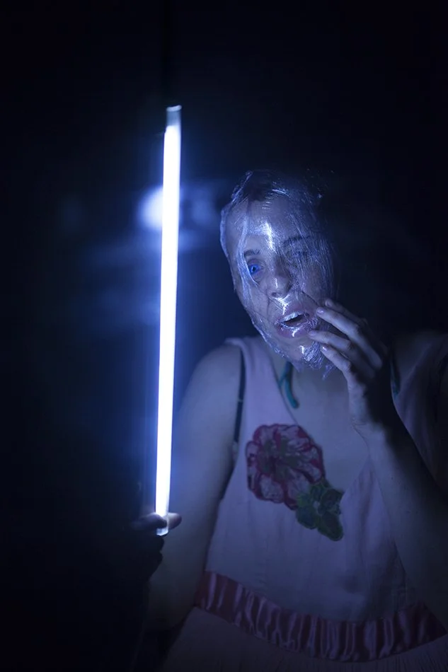 A woman with a transparent face mask glows in the dark while holding a vertical blue light stick, with an expression of surprise or amazement.