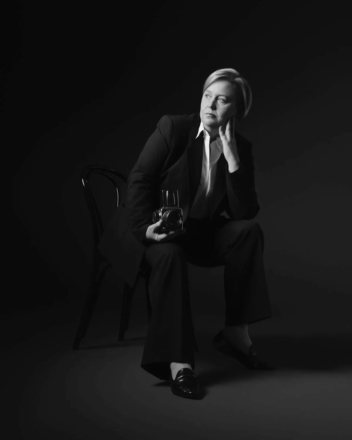Black and white photo of a woman sitting on a chair, holding a vintage camera, with a contemplative expression, looking to the side, in a dark studio setting.