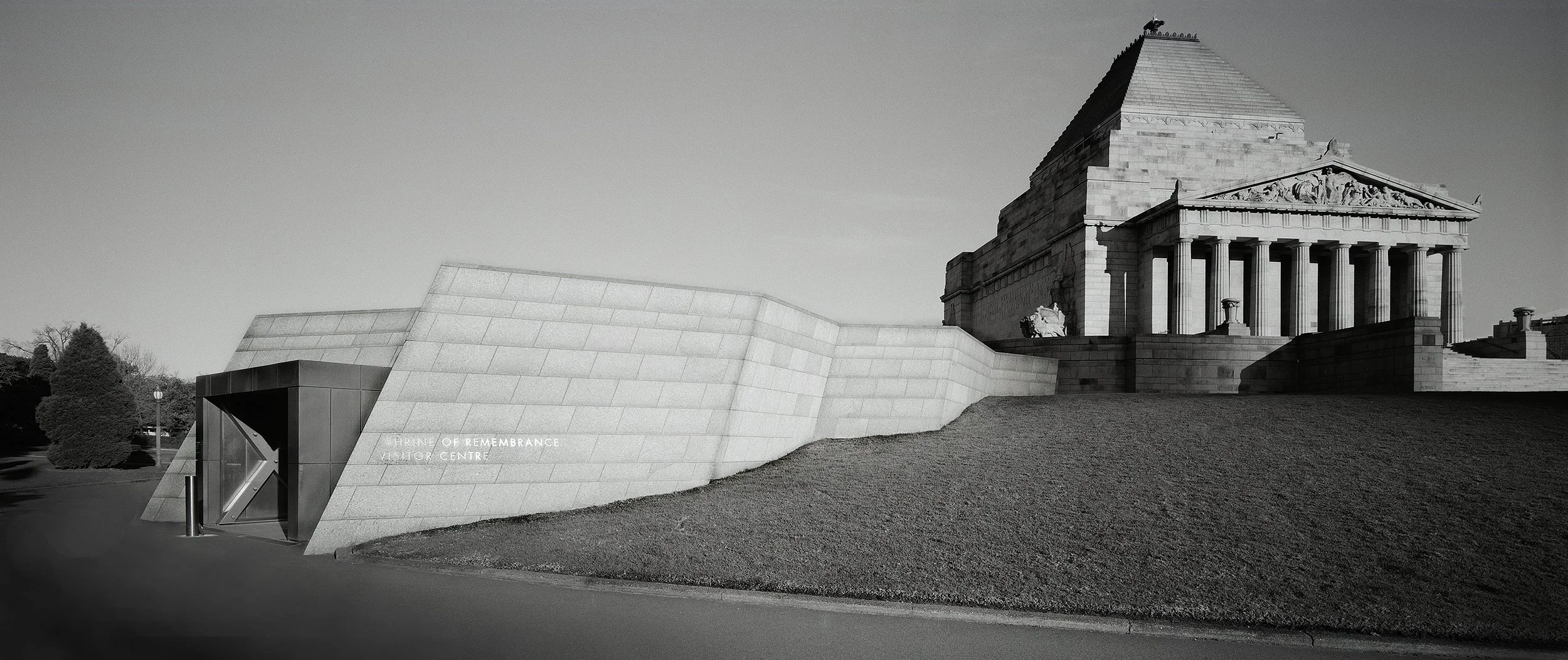 Black and white photograph of the National War Memorial, a large memorial with a classical temple design with columns, situated on a raised platform with stairs. To the left, a modern visitor center with angular design and inscription 'URNE OF REMEMBRANCE VISITOR CENTRE' is visible. There are trees and a pathway surrounding the memorial.
